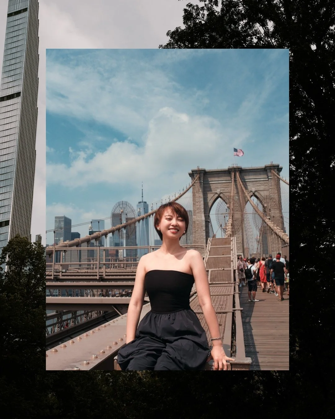 A woman in a black strapless top and black pants smiling while sitting on a ledge of the Brooklyn Bridge in New York City. The bridge and city skyline with tall buildings and a partly cloudy sky are visible in the background.