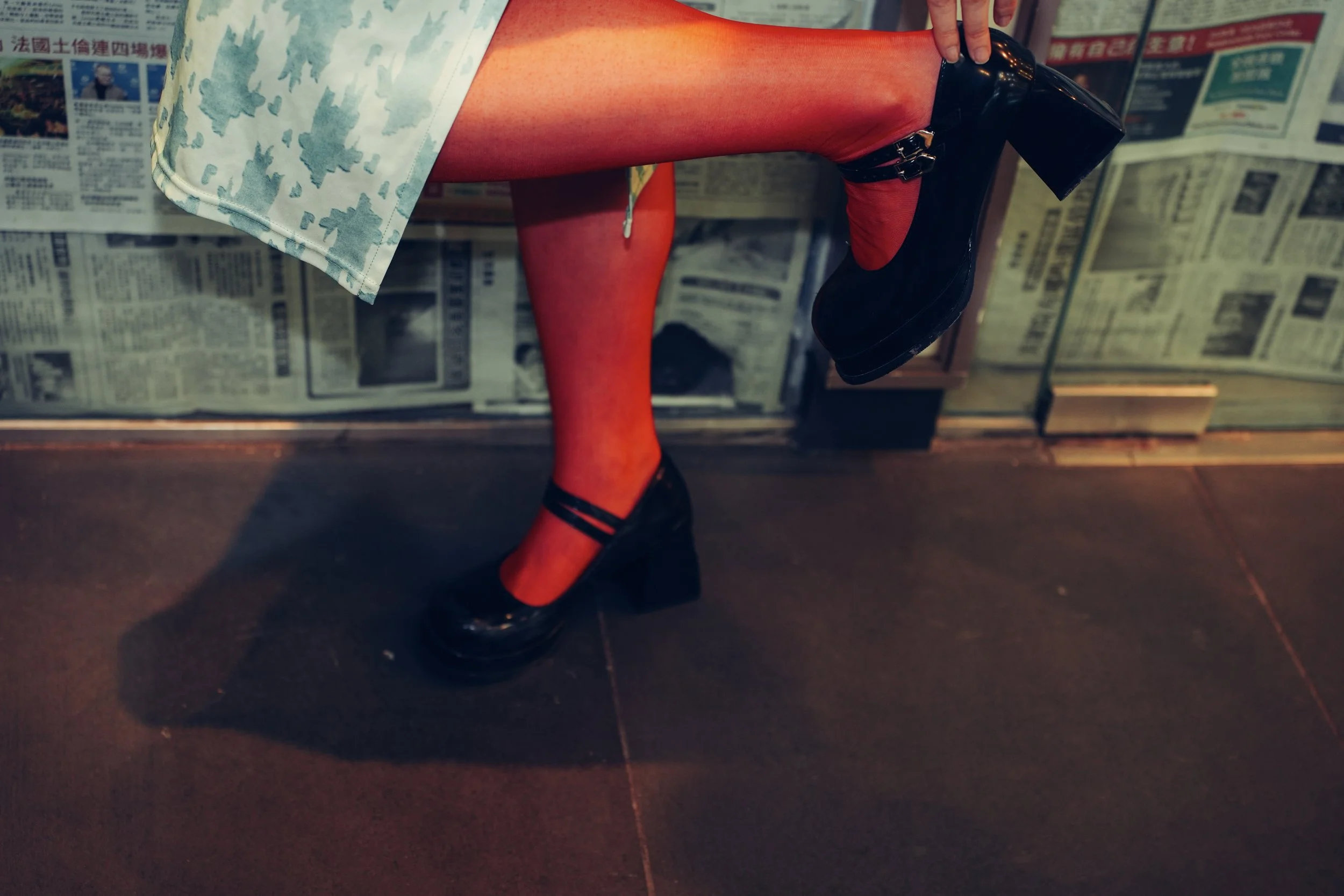 Close-up of a person wearing a blue patterned skirt, red tights, and black high-heeled shoes, standing on a surface with newspaper-covered walls in the background.