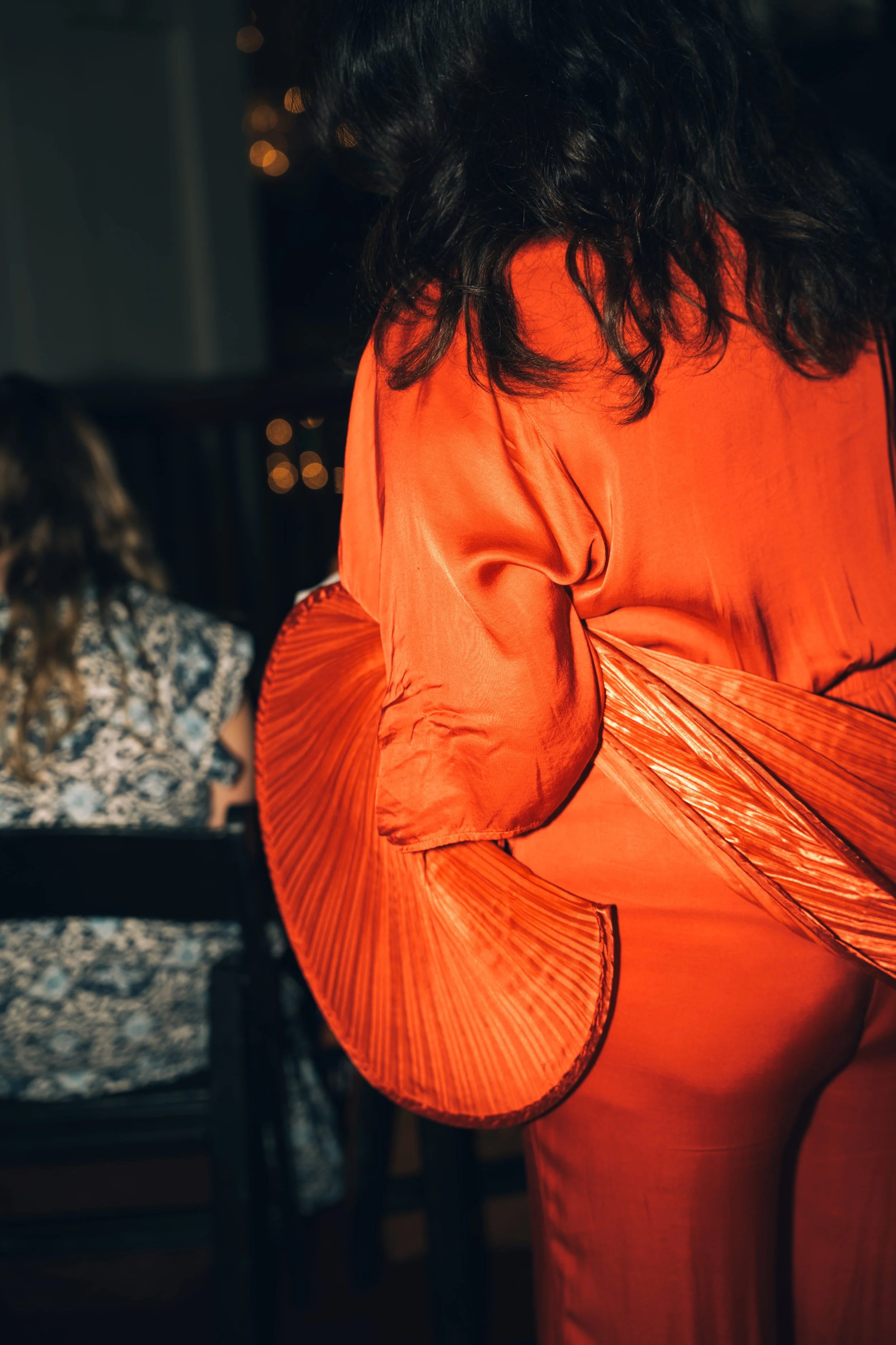A person wearing an orange dress with pleated ruffles on the long sleeve, with black curly hair. In the background, there are hanging string lights and a woman with long wavy hair wearing a gray patterned top, sitting at a table.