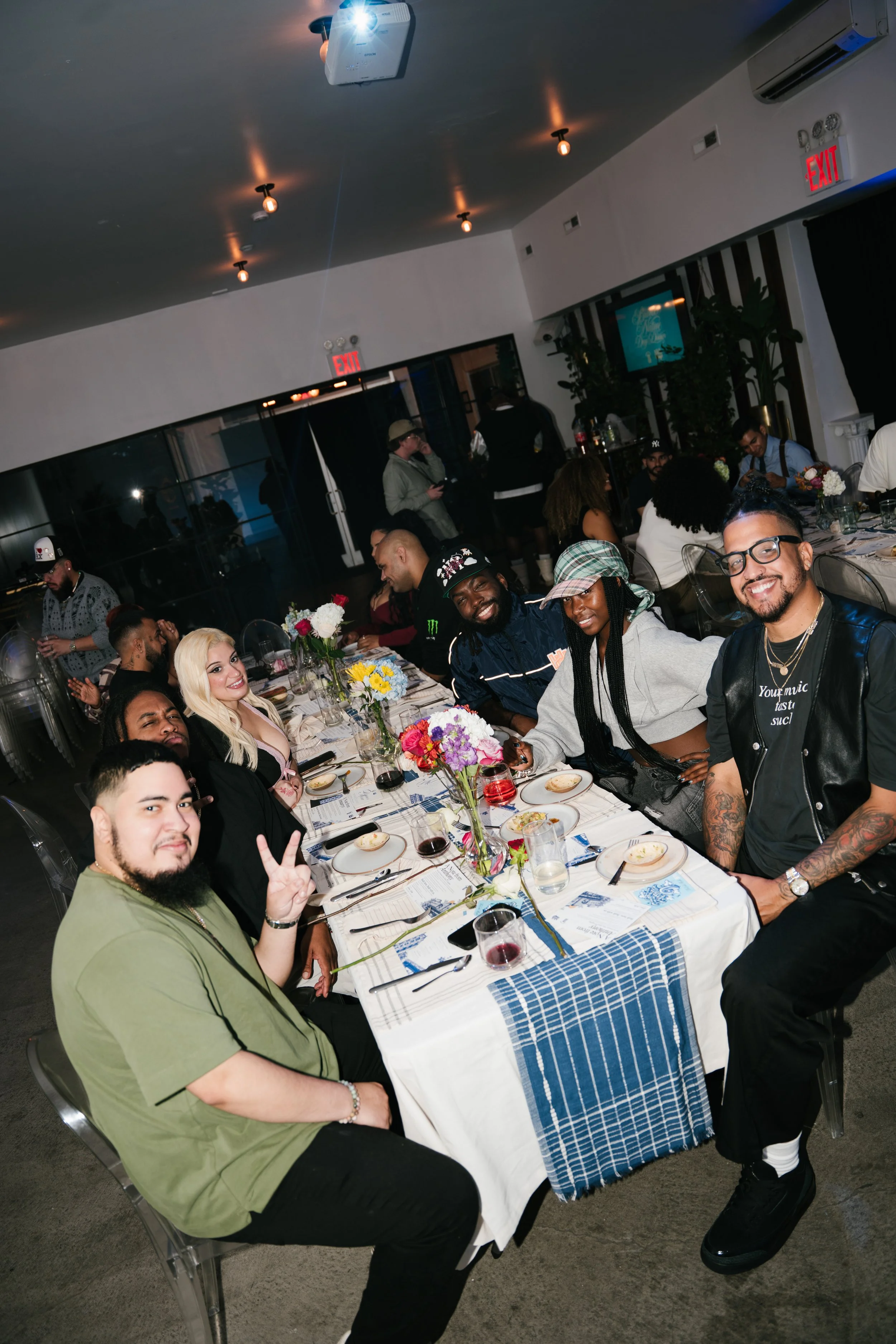 People sitting around a dining table at a celebration or event, smiling and enjoying food, with a floral centerpiece, set in a modern restaurant or banquet hall.