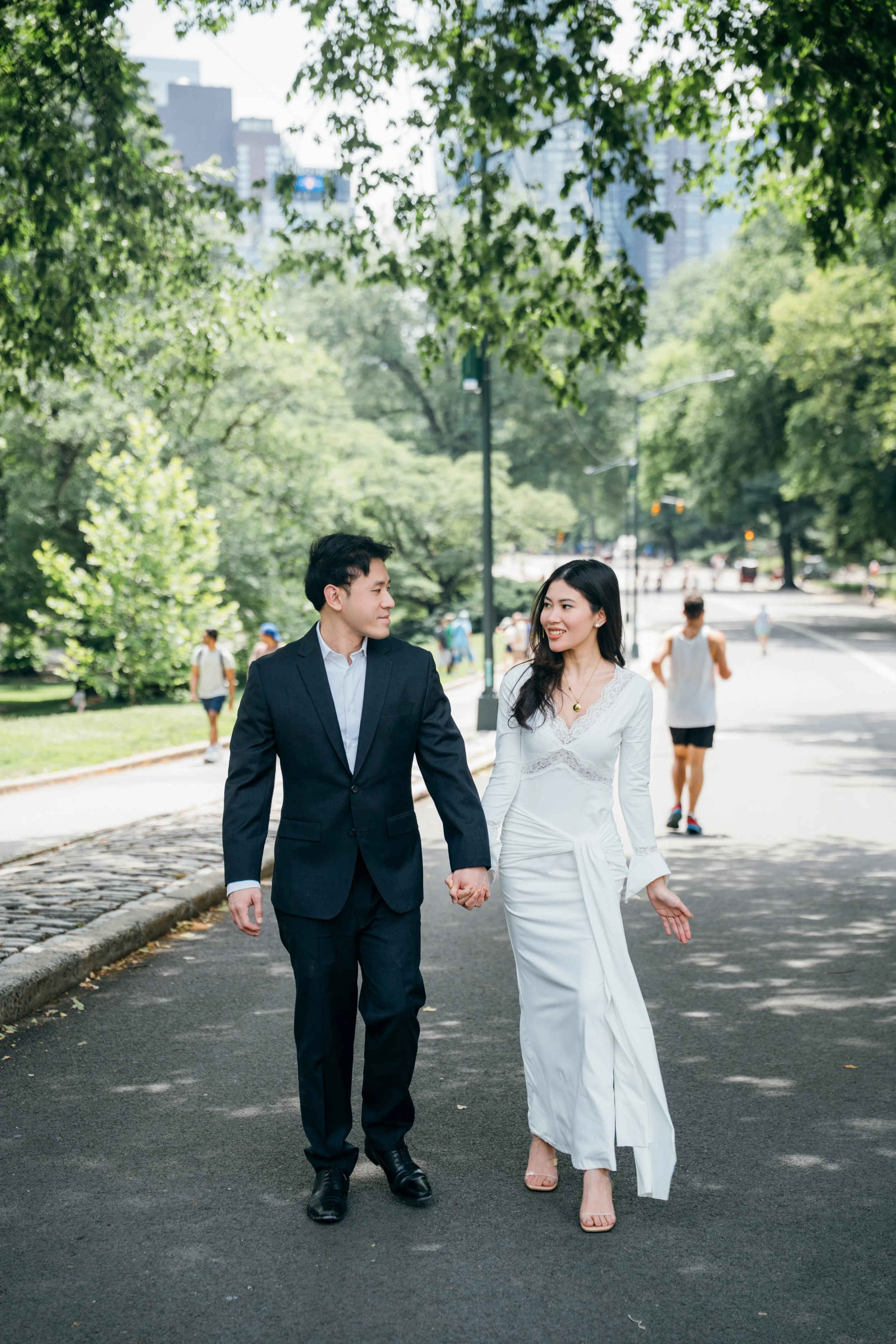 A couple walking hand in hand through a city park on a sunny day, holding hands and smiling at each other, with other people jogging and walking in the background.