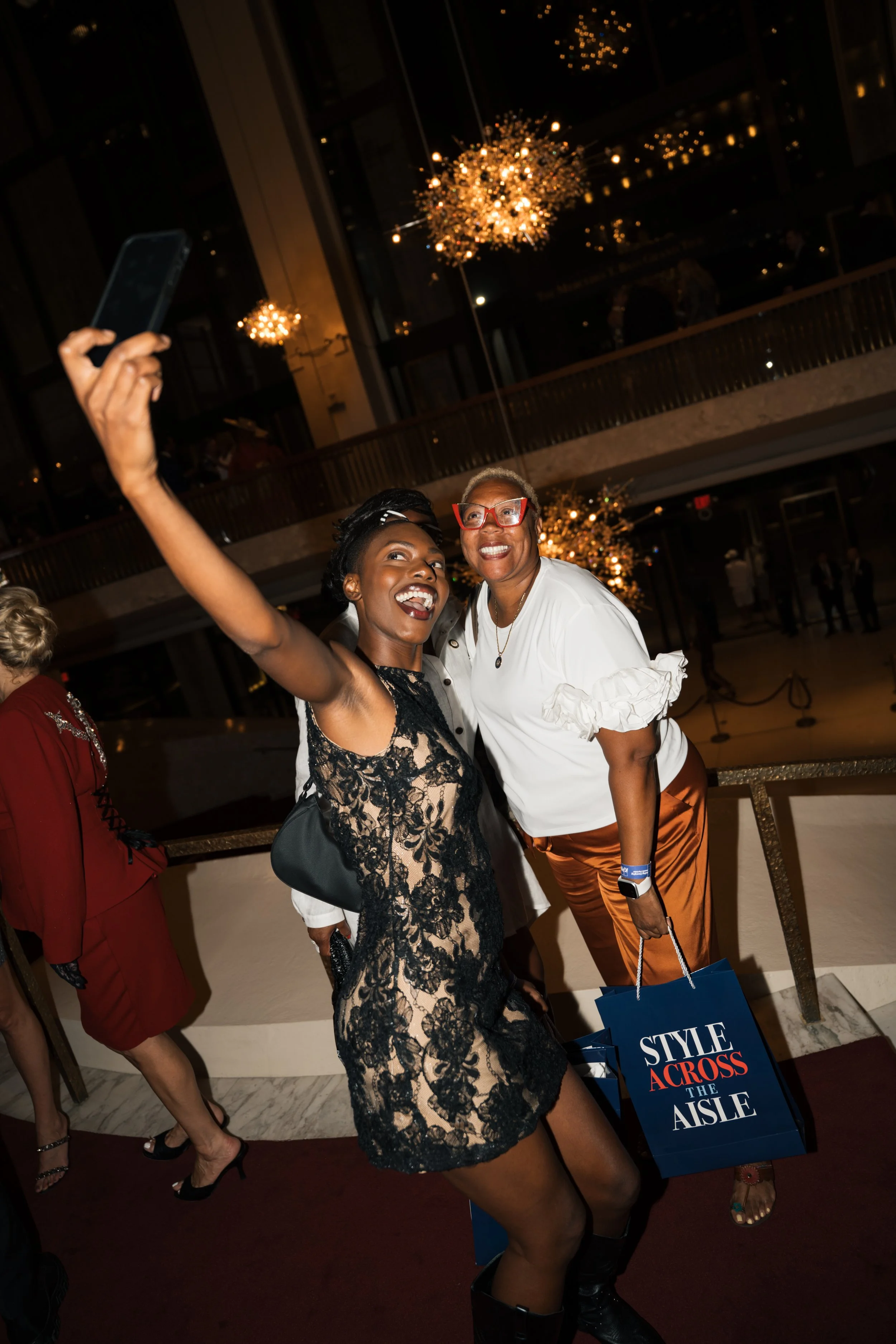 Two women taking a selfie at an indoor event, with chandeliers and other people in the background. One woman is wearing a black lace dress, and the other is wearing glasses and a white blouse, holding shopping bags. They are smiling and posing for th