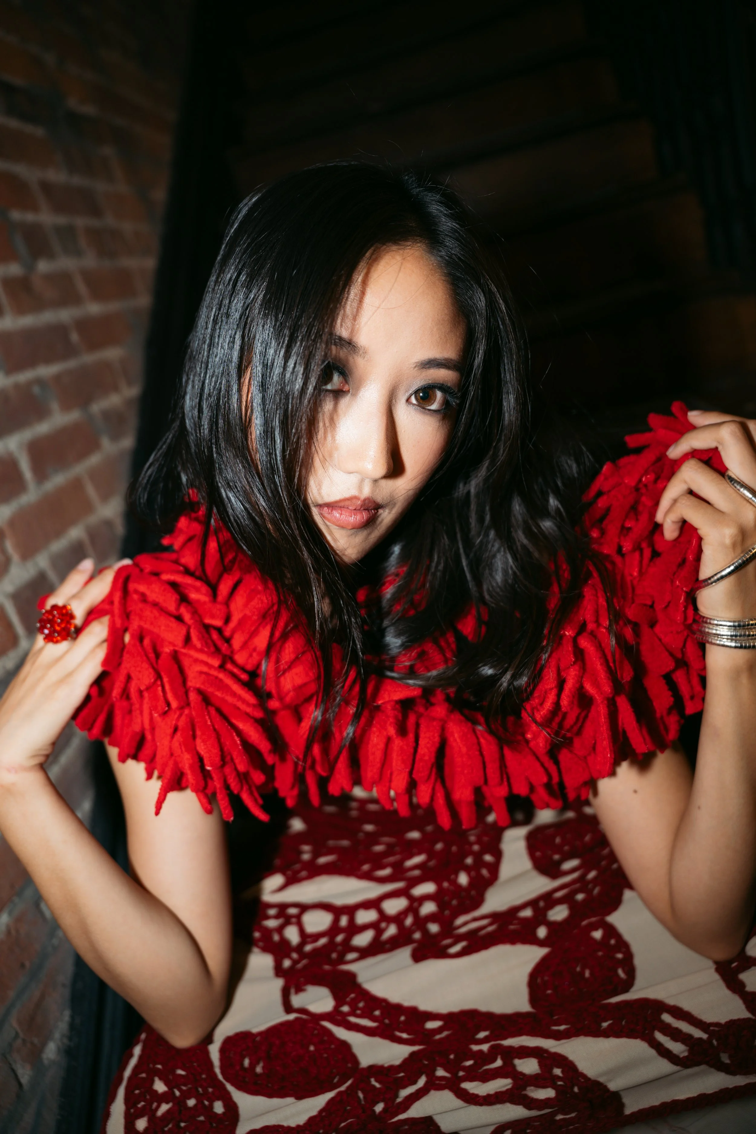 A woman with long dark hair, wearing a red fringed top, is looking at the camera with a serious expression. She is holding her hair with one hand and is sitting in front of a brick wall.