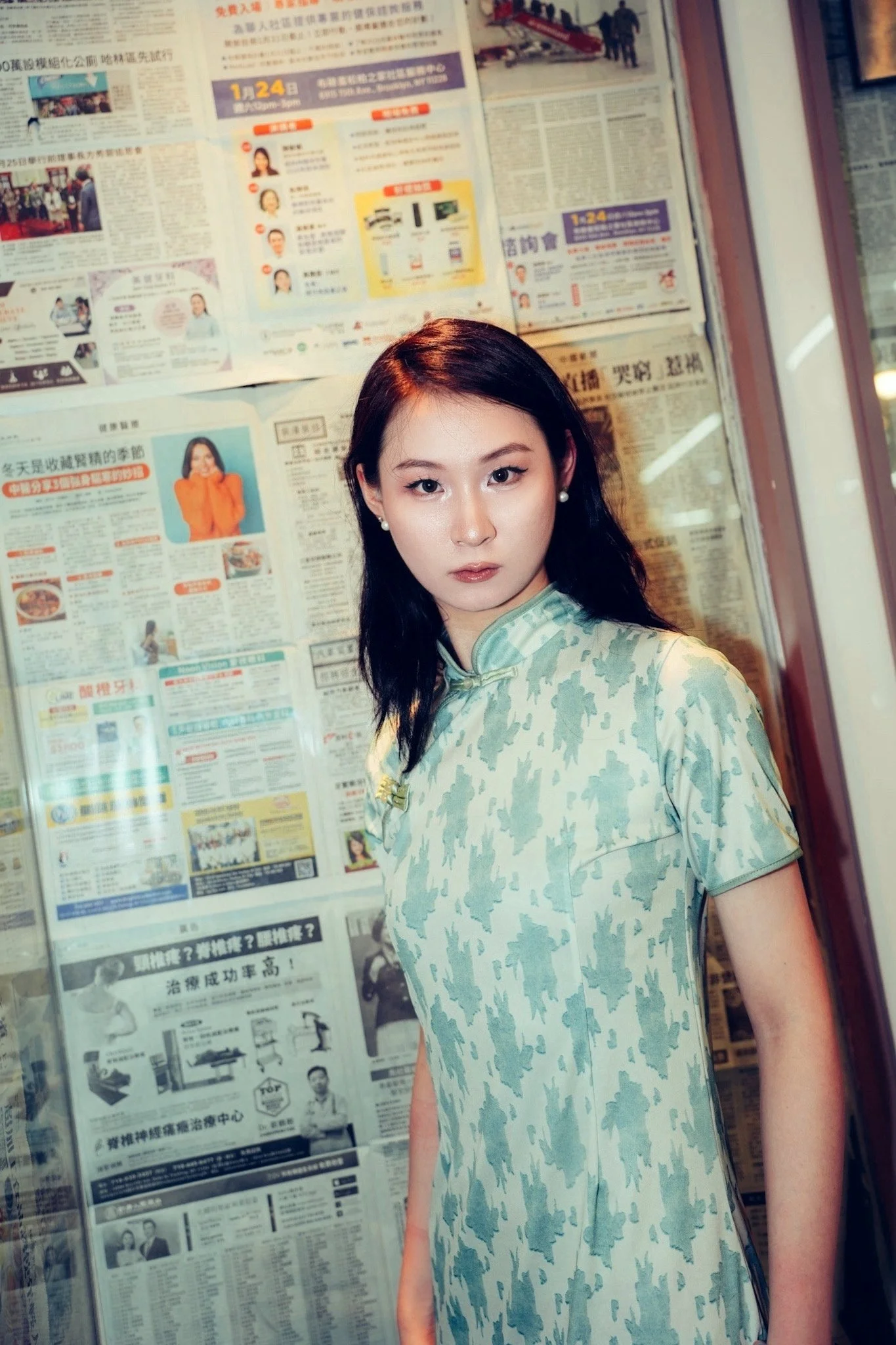 A young woman with black hair, wearing pearl earrings and a green camouflage dress, stands in front of a wall covered with newspapers in Chinese.