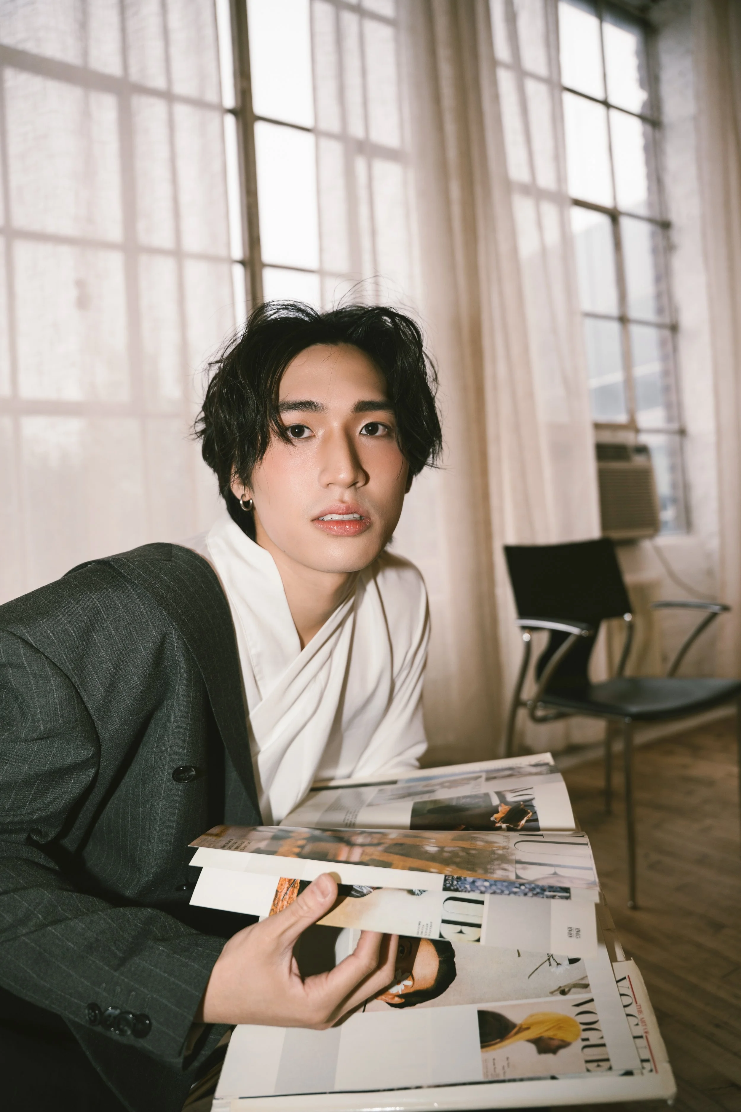 A person with dark, wavy hair, wearing a dark pinstripe suit and a white shirt, is sitting at a table in an urban-style room with large windows, looking directly at the camera. They are holding open a magazine with fashion photos, and there are chair