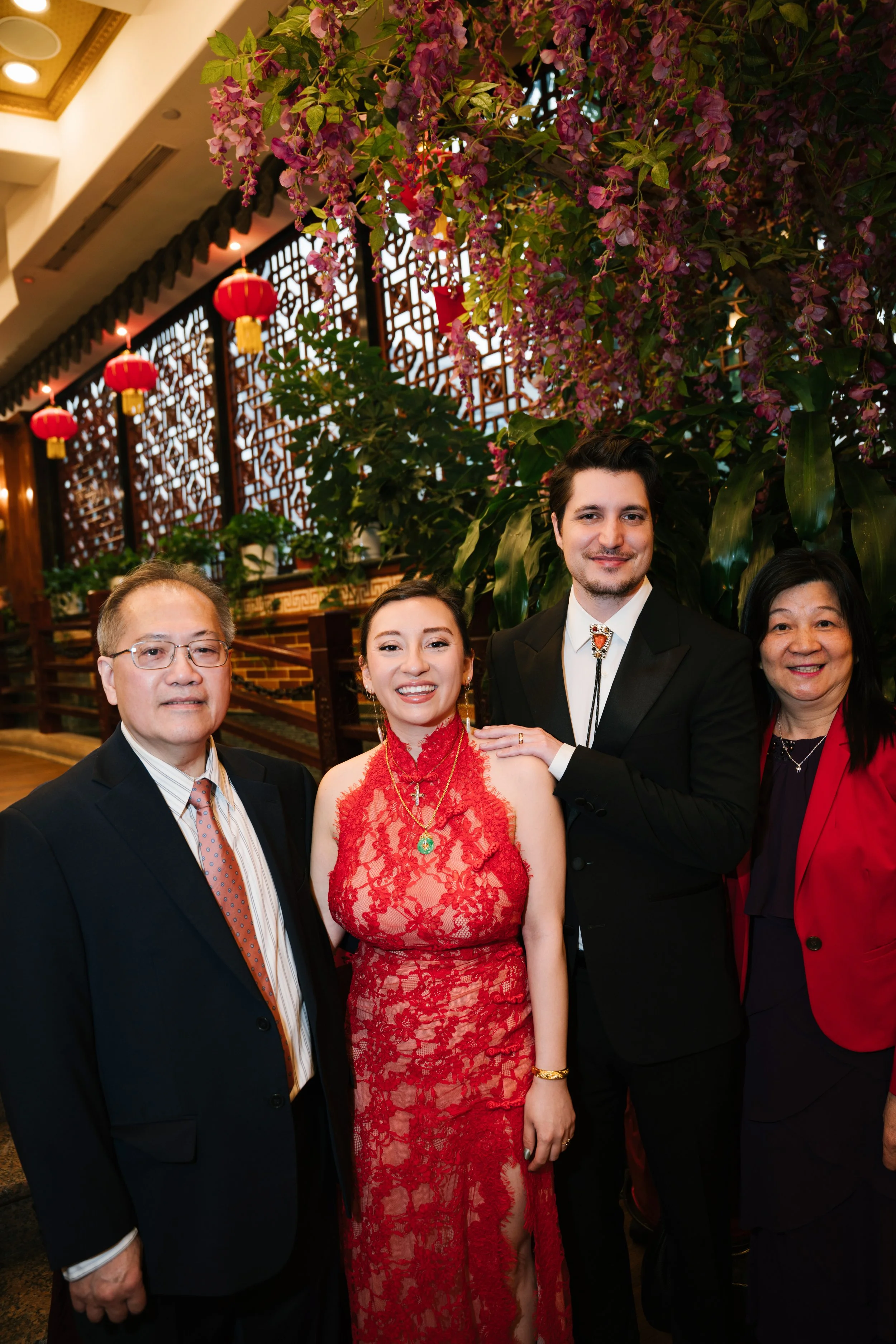 Group of people dressed in formal attire standing in a decorated interior with pink flowering plants and red lanterns.
