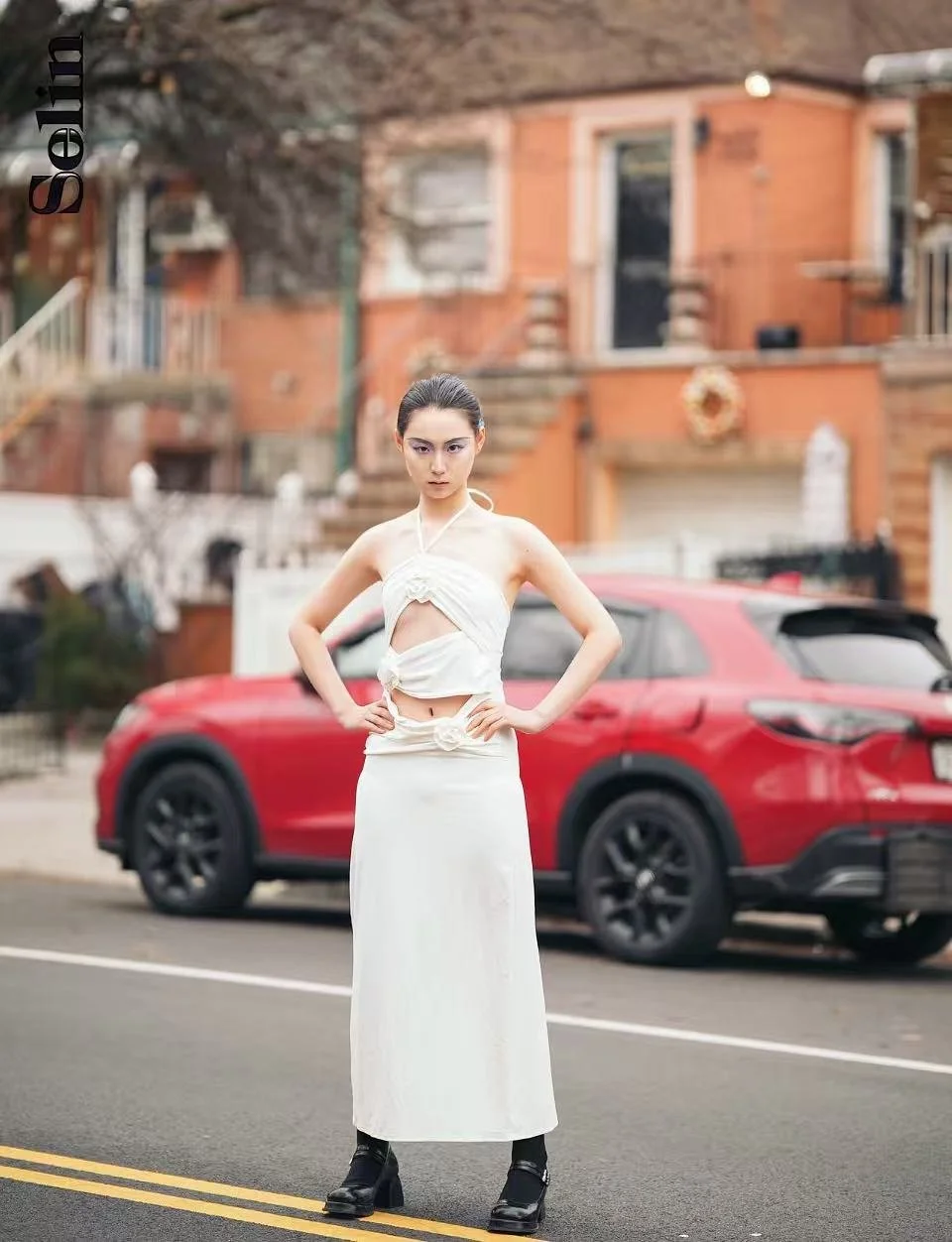 A woman in a stylish white dress standing confidently on a street with a red SUV parked behind her, in front of reddish-brown houses.