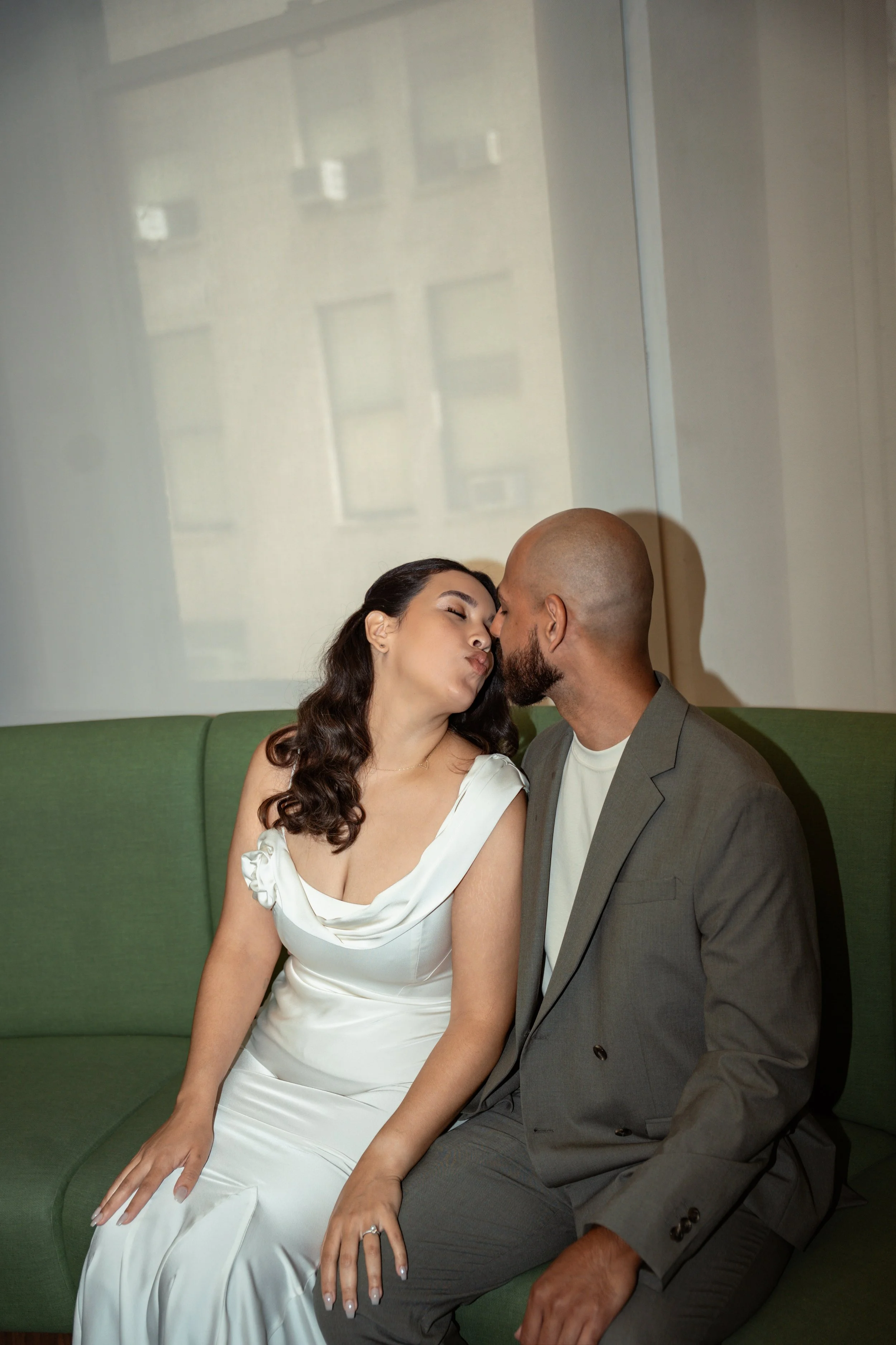A woman in a white dress and a man in a gray suit sit close together on a green sofa, sharing a tender moment with their foreheads touching and eyes closed in a cozy, intimate setting.