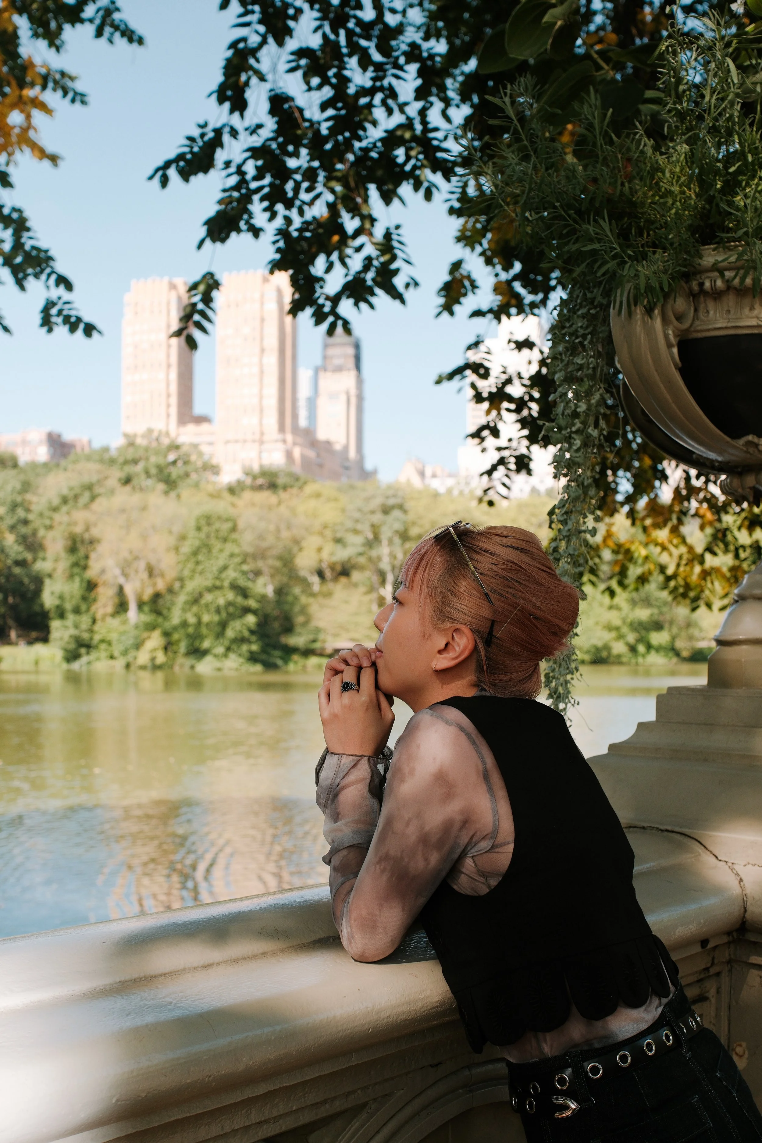 A woman with reddish-brown hair and tattoos on her arm, standing by a river in Central Park, New York City, with tall buildings in the background. She is leaning on a stone railing, her hands clasped in front of her face in a prayer or contemplative 