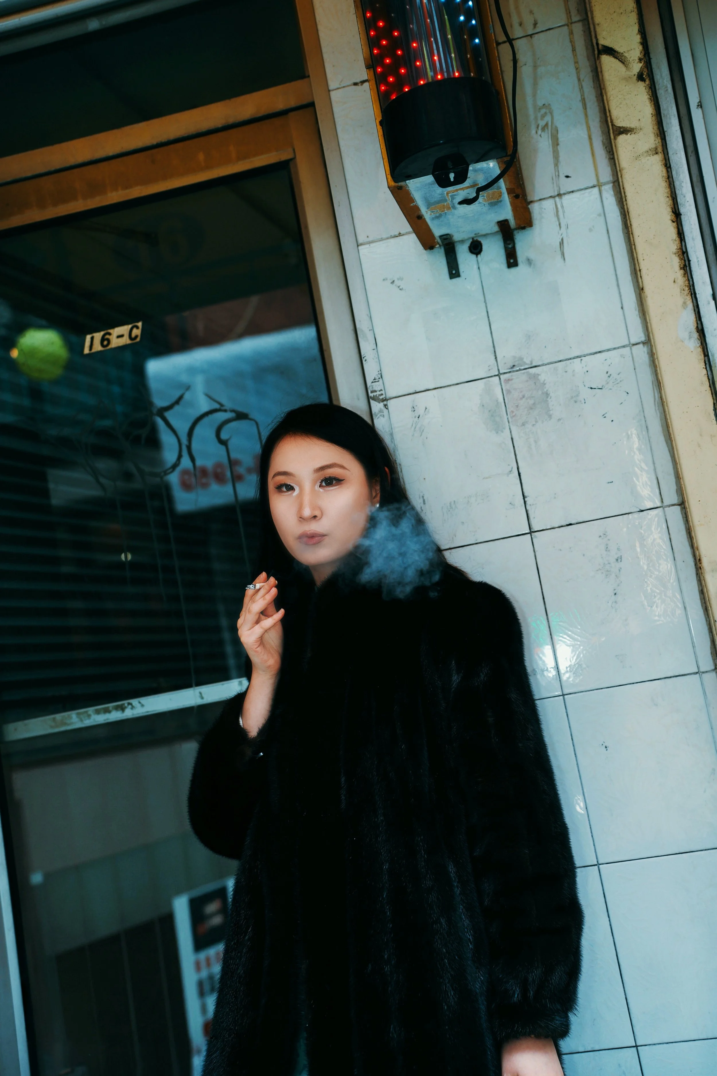 A woman with black hair wearing a black fur coat, smoking with smoke visible in front of her face, standing in front of a tiled wall next to an elevator with a red and blue light panel.