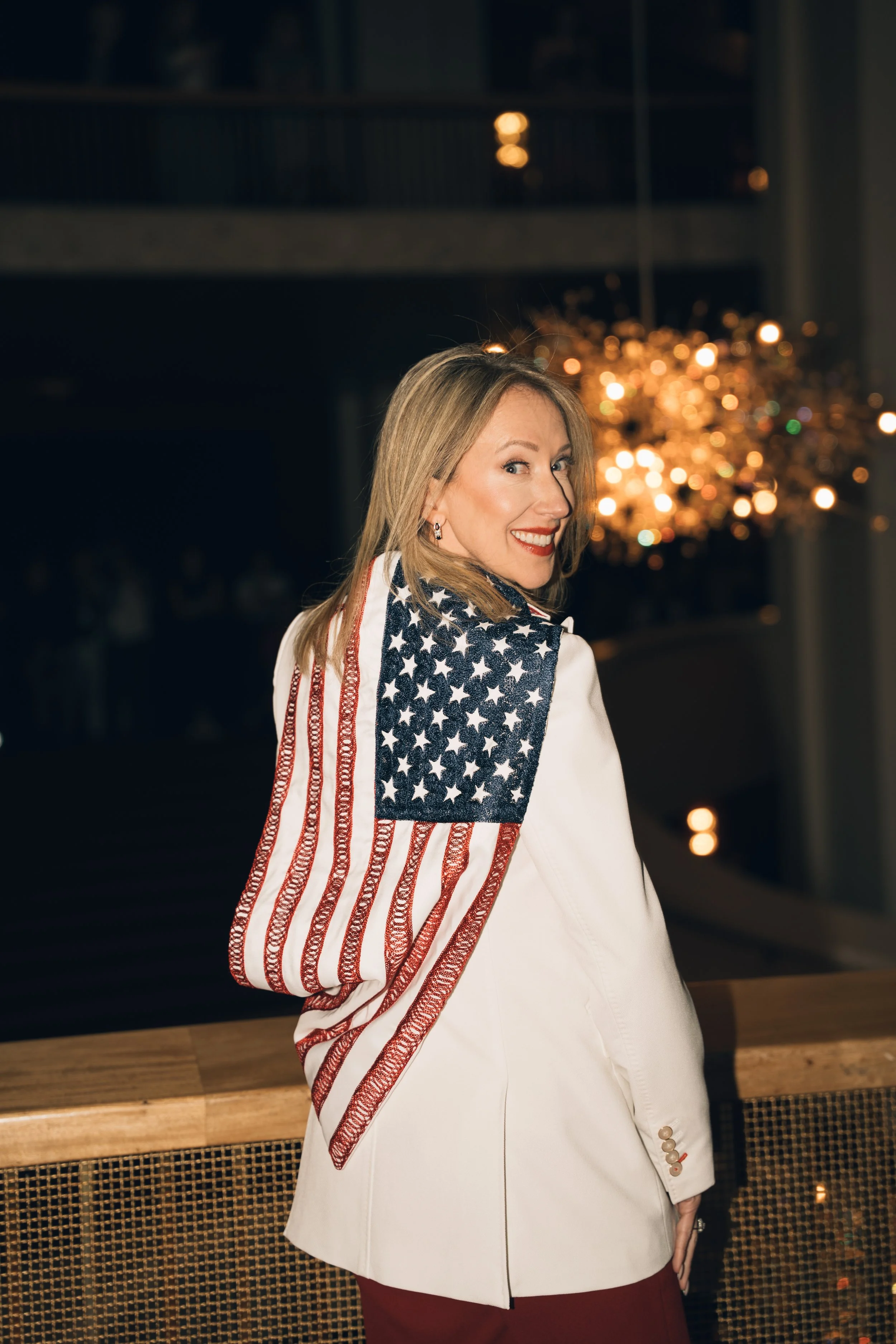 A woman with blonde hair, wearing a white blazer with star-spangled accents on the sleeve, standing indoors with a blurred string of lights in the background.