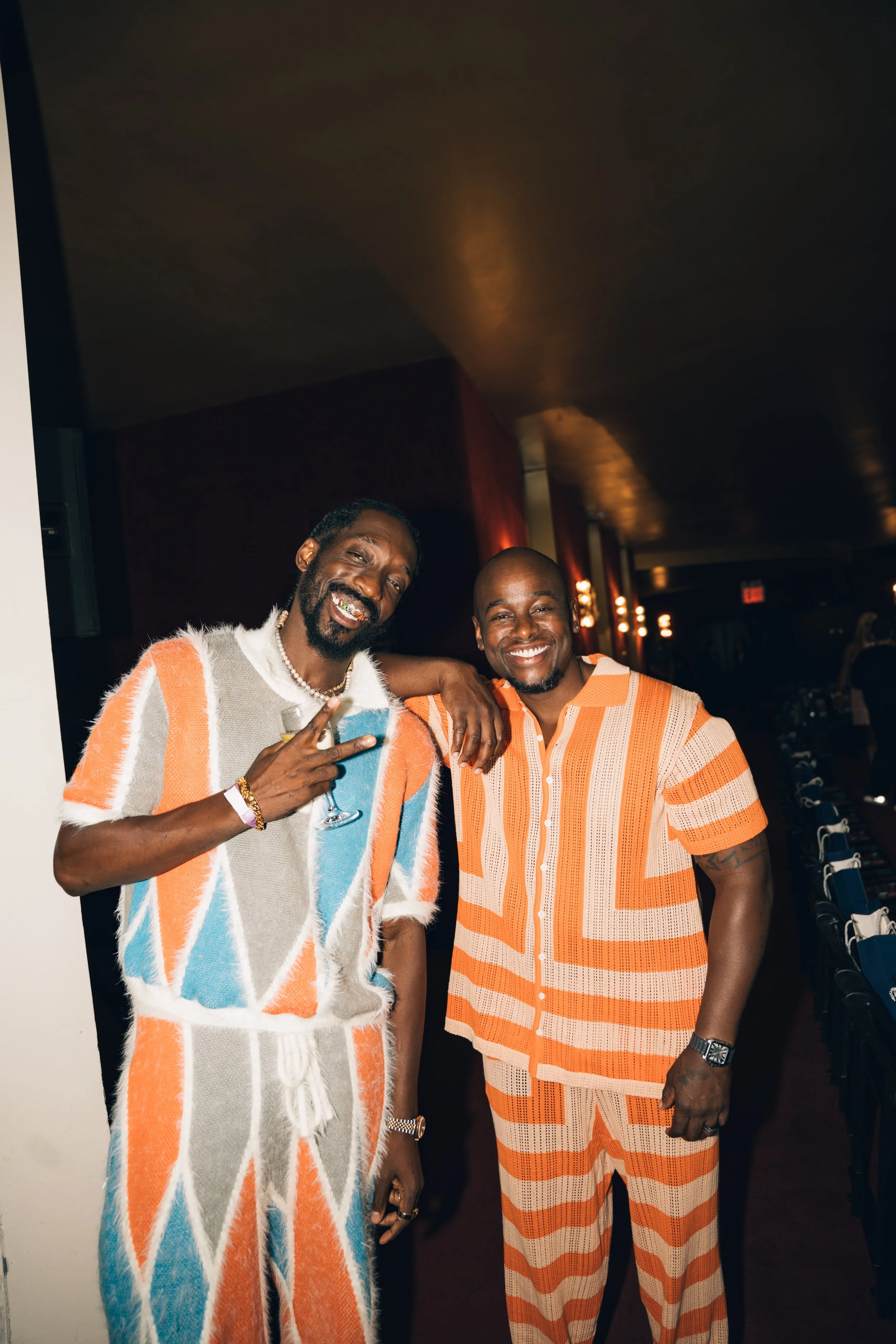 Two men are smiling and posing together at an indoor event. The man on the left is wearing a colorful, striped shirt with orange, white, blue, and beige patterns, and has a beard with gold teeth. The man on the right is wearing an orange and beige st
