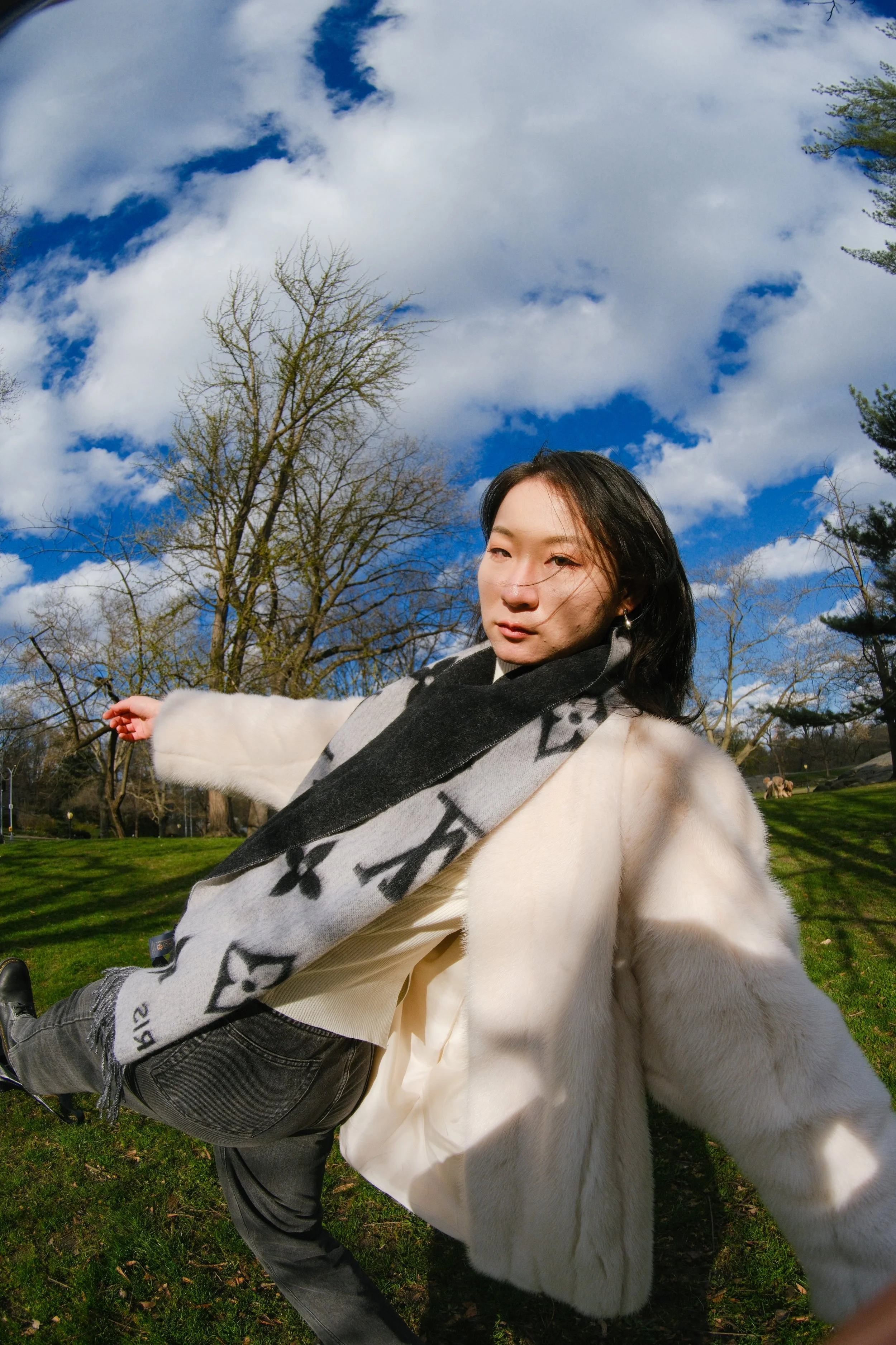 A woman with long dark hair wearing a white coat and a black and white scarf, standing outdoors in a park with trees and grass, under a blue sky with clouds.