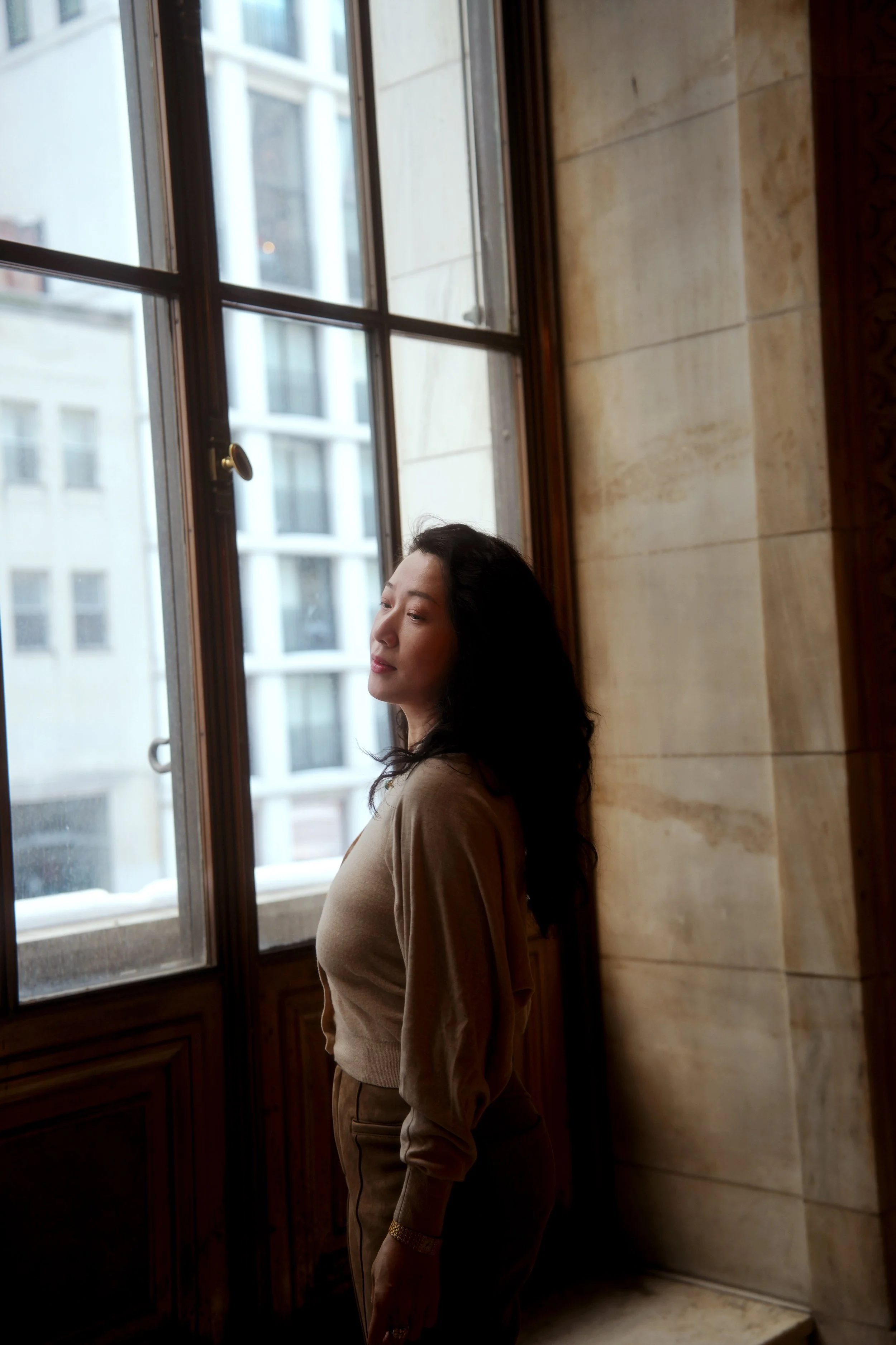 A woman standing by a large window with wooden framing, looking contemplative with her eyes closed, in an interior space with marble walls.