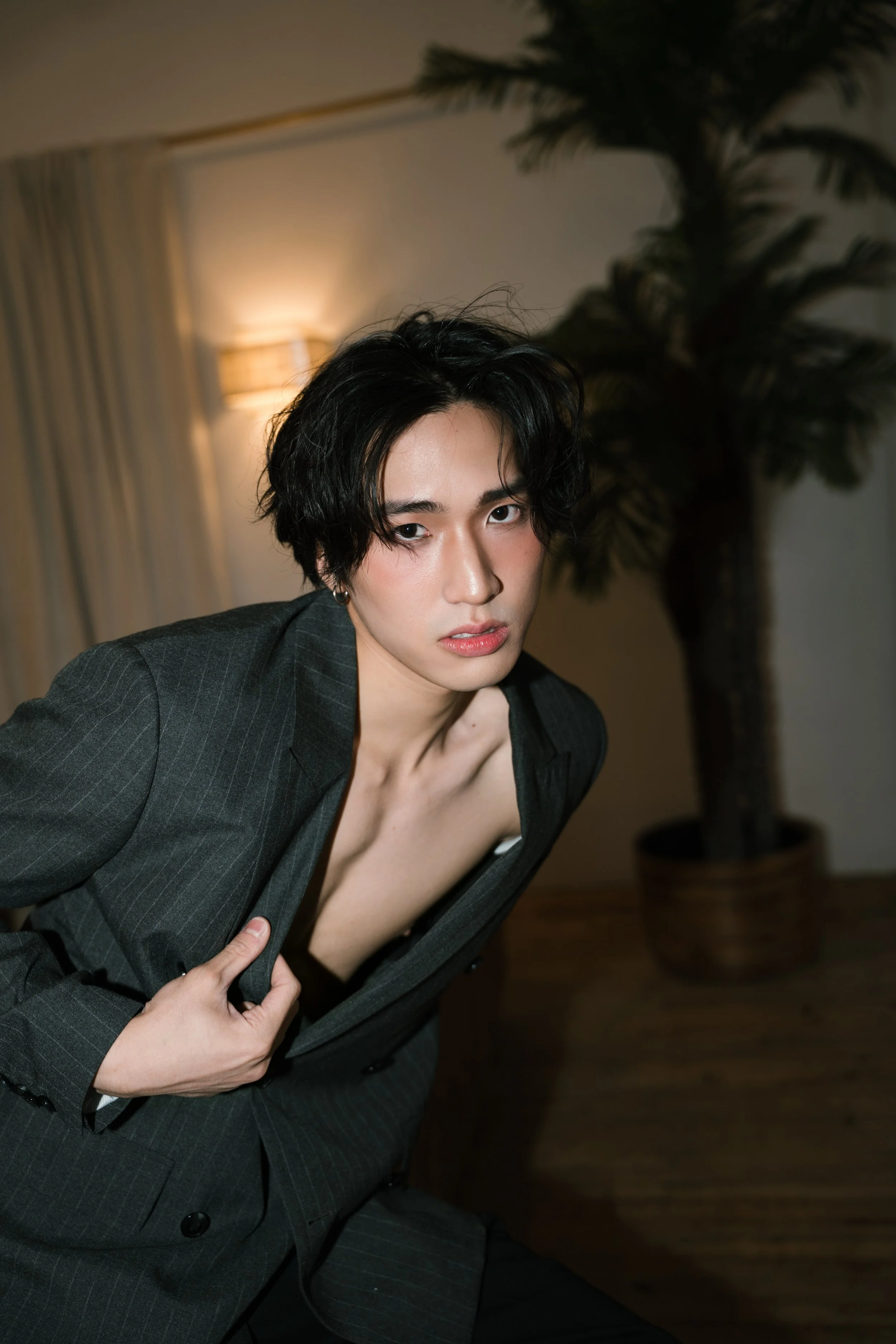 A person with short, curly black hair and makeup, wearing a dark blazer with a deep neckline, is seated indoors with a potted plant and a lamp visible in the background.