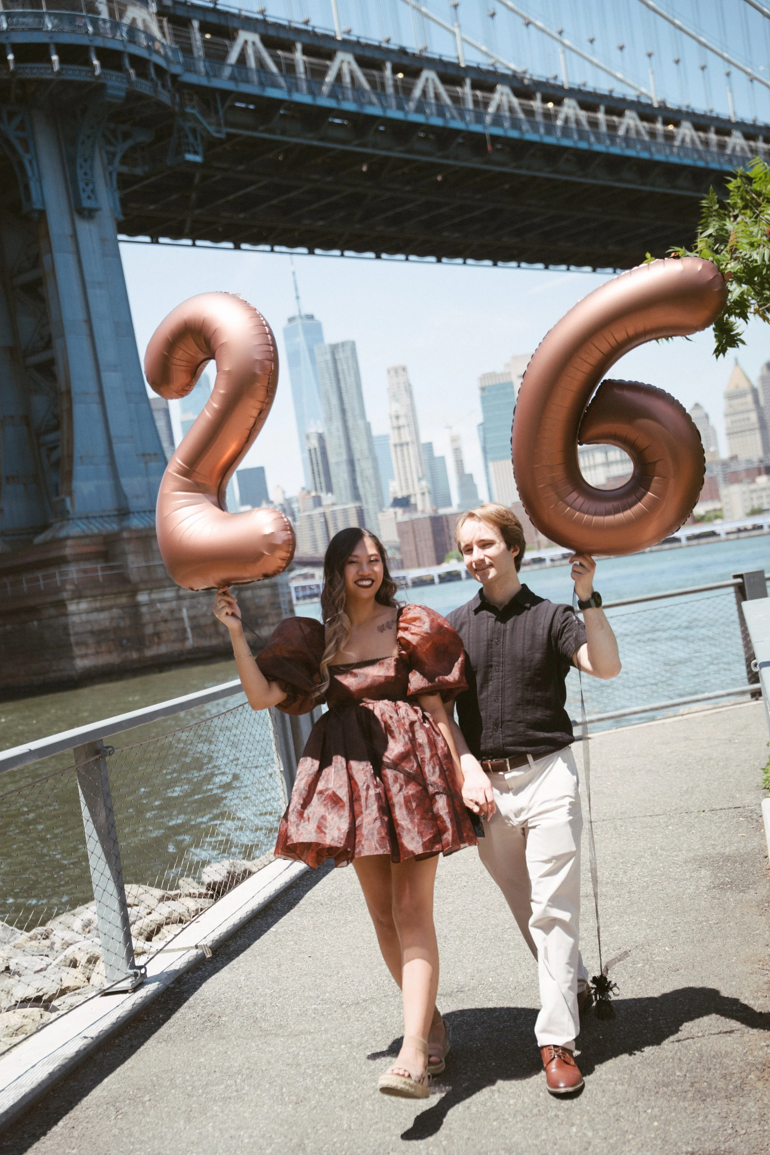 Two people, a woman and a man, walking on a bridge near water with a city skyline in the background. The woman is holding balloon numbers '2' and '6'.