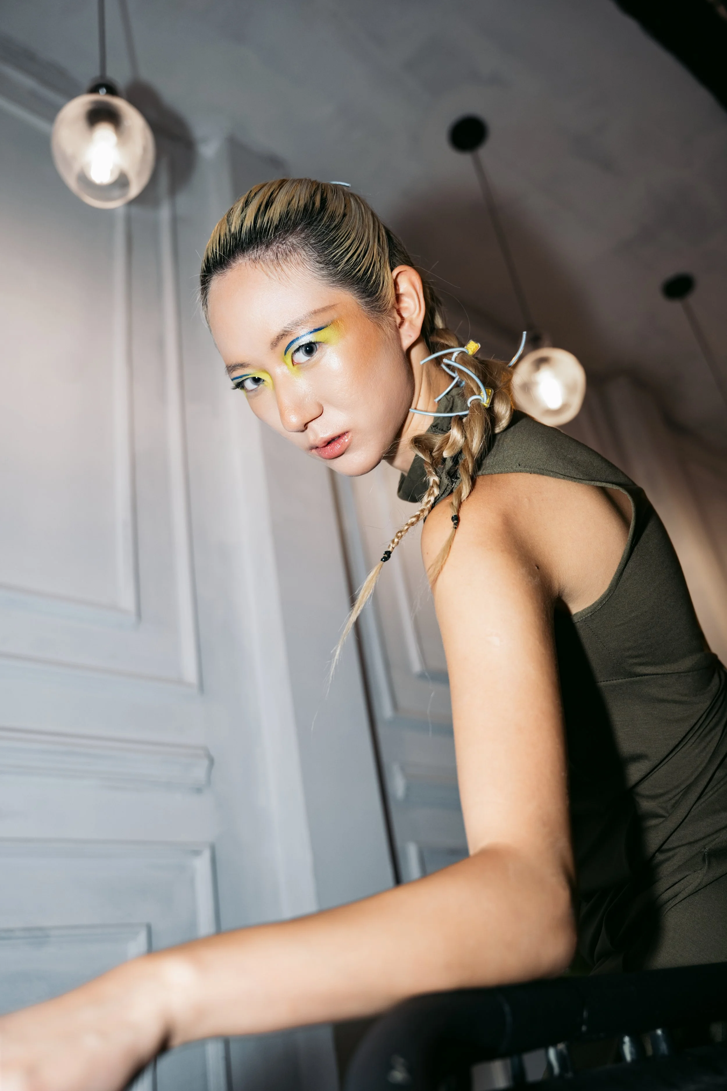 A woman with braided hair and bright makeup, wearing a dark sleeveless top, seated indoors, with hanging ceiling lights in the background.