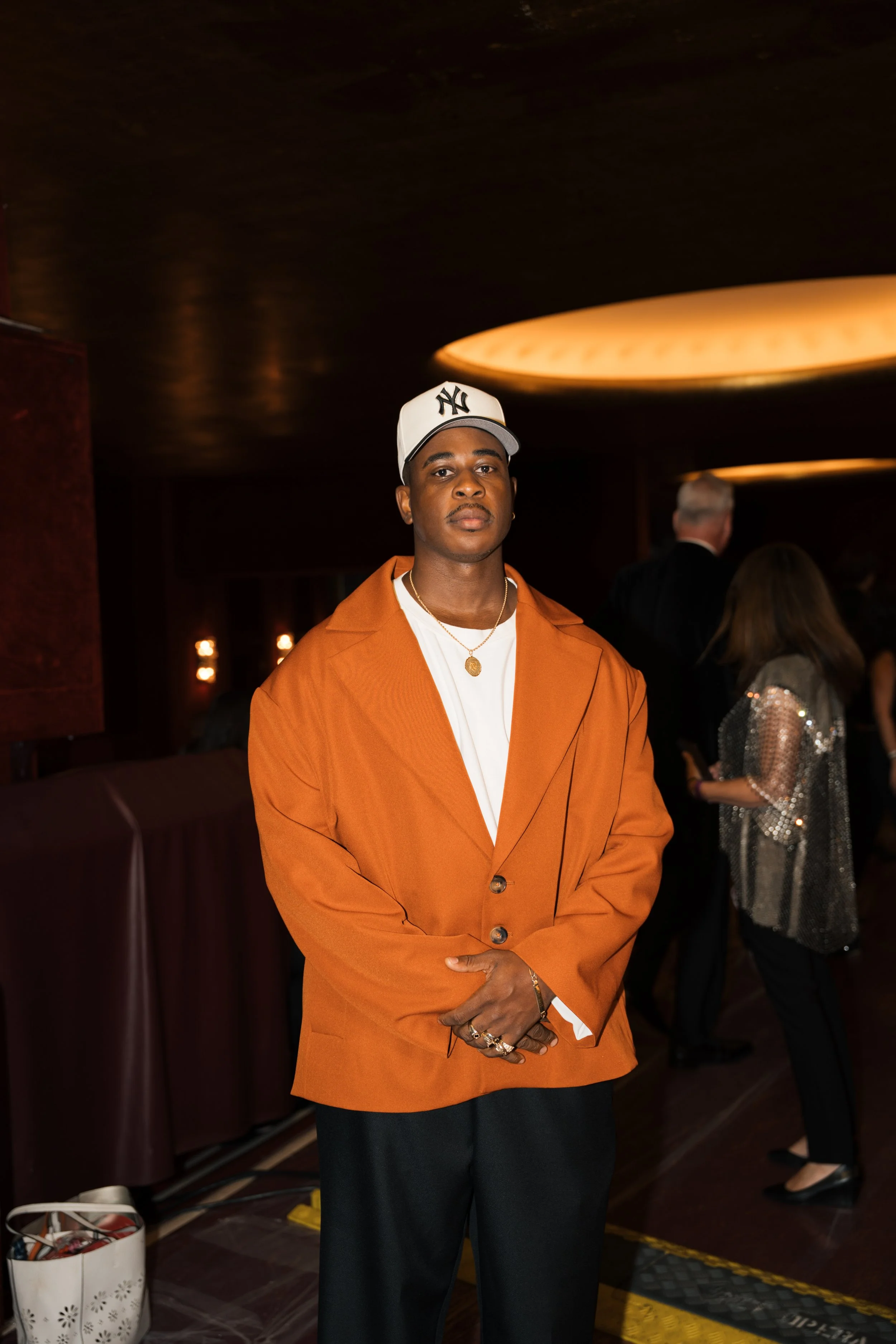 A man wearing a white New York Yankees baseball cap, an orange blazer, and black pants standing in a dimly lit room with a dark ceiling and a warm-colored overhead light. There are people in the background, including a woman in a shiny dress and an o