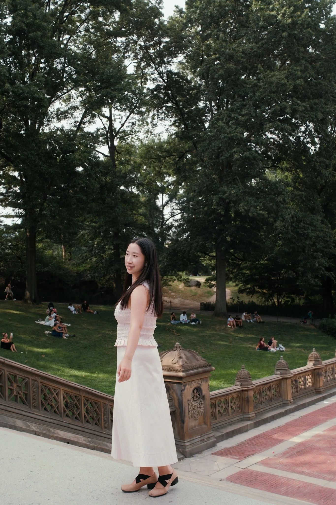 A young woman in a light pink top and long white skirt stands on a bridge with ornate railings, looking to her right. In the background, a park with large trees and people sitting on the grass can be seen.