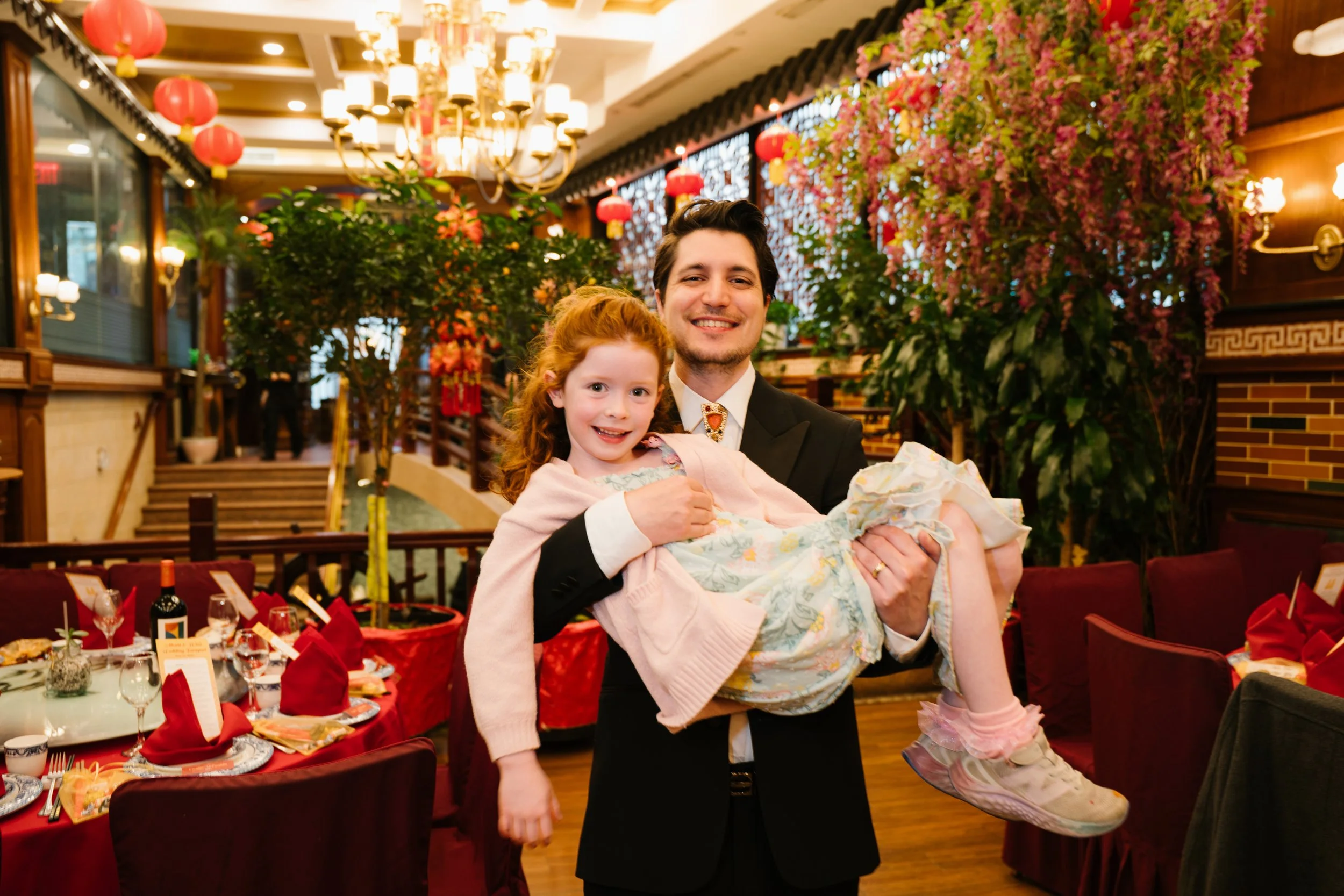 A man in a tuxedo holding a smiling young girl in a pink sweater at a decorated banquet hall with red lanterns, flowers, and tables set for a celebration.