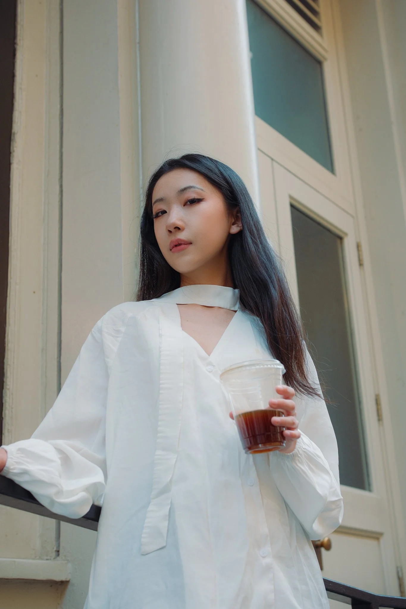 A woman with long dark hair, wearing a white blouse, holds a plastic cup filled with iced coffee or tea while standing outdoors near a beige building with large windows.