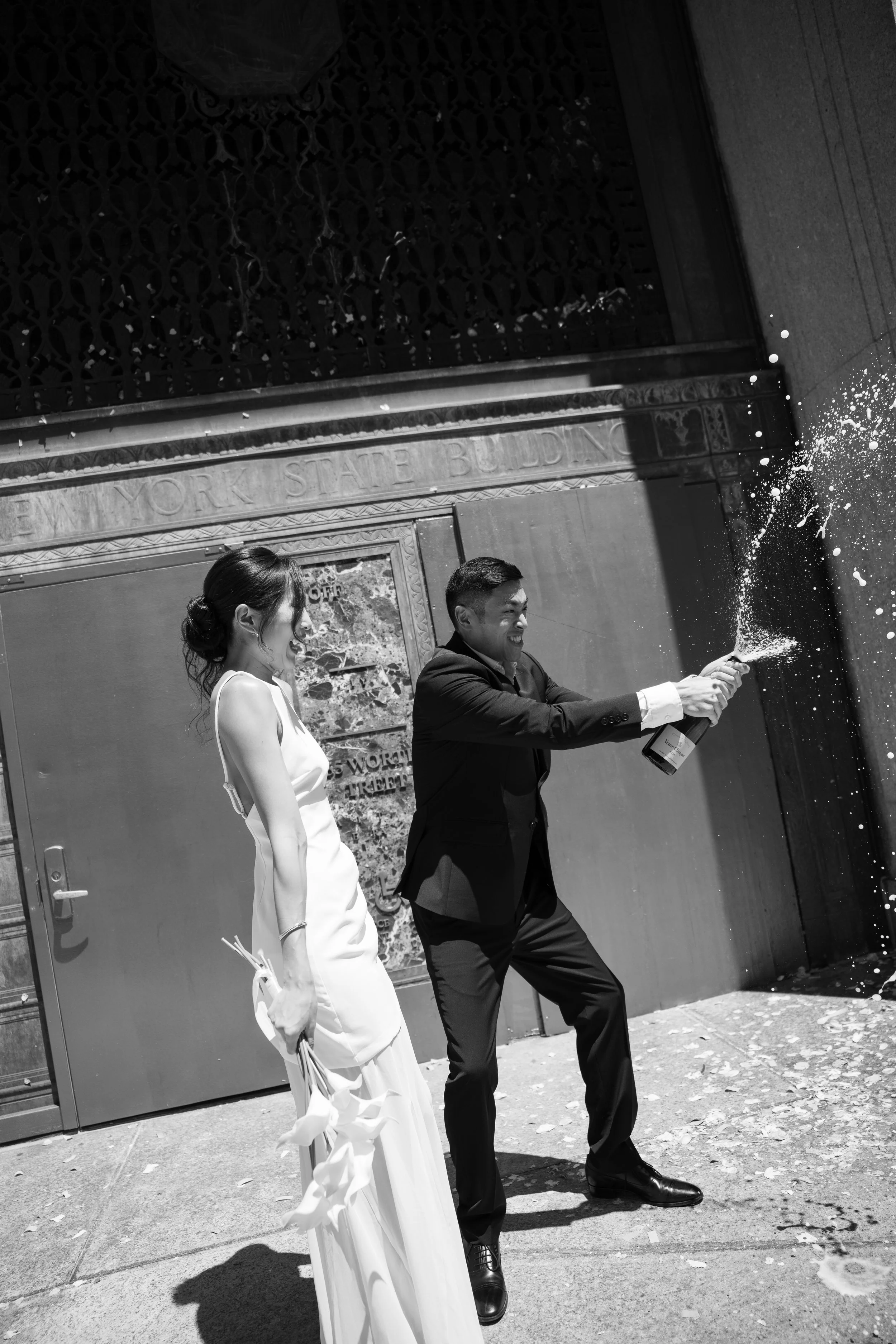 Two people, a woman in a white dress holding flowers and a man in a suit, celebrating by popping a bottle of champagne on a city sidewalk in front of a historic building.