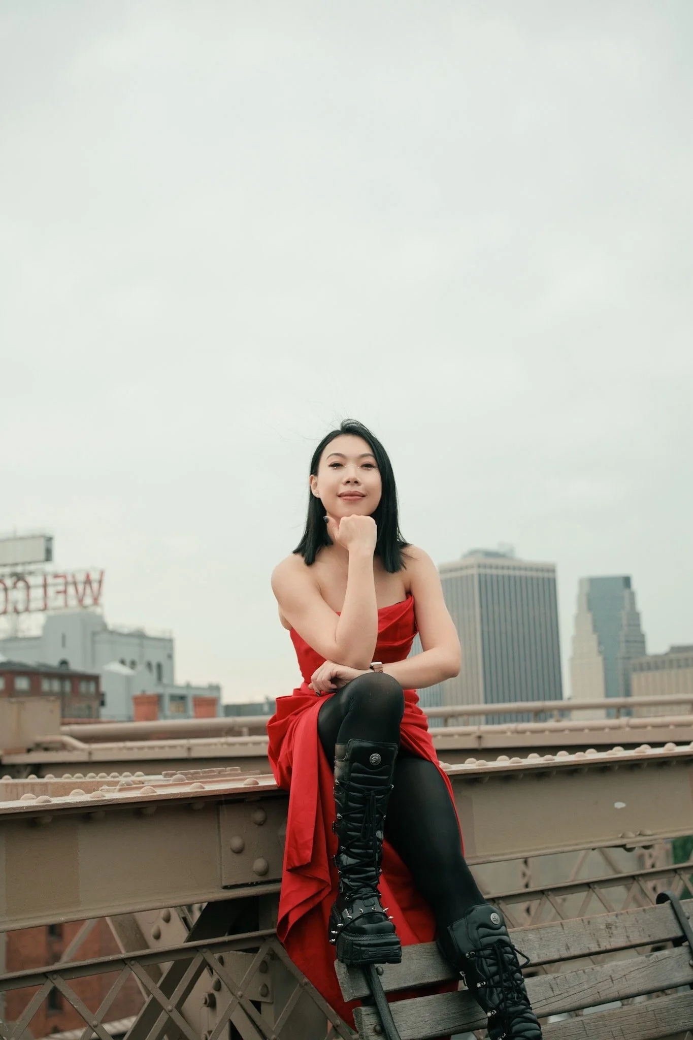 A woman in a red dress and black boots sitting on a bridge with a cityscape in the background.