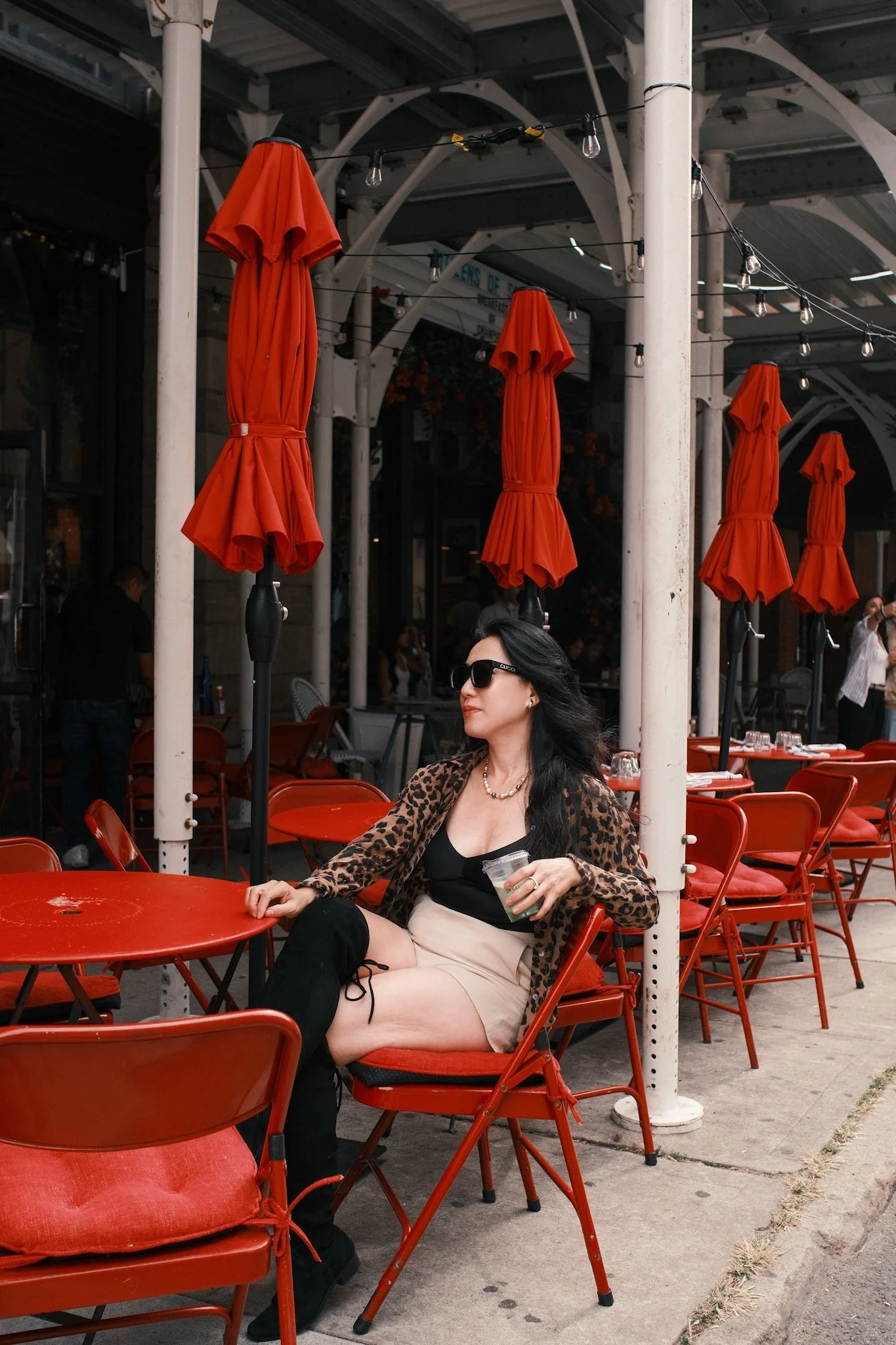 A woman sitting at an outdoor cafe table with red chairs and orange umbrellas, holding a drink and wearing sunglasses, a leopard print jacket, and a black top.