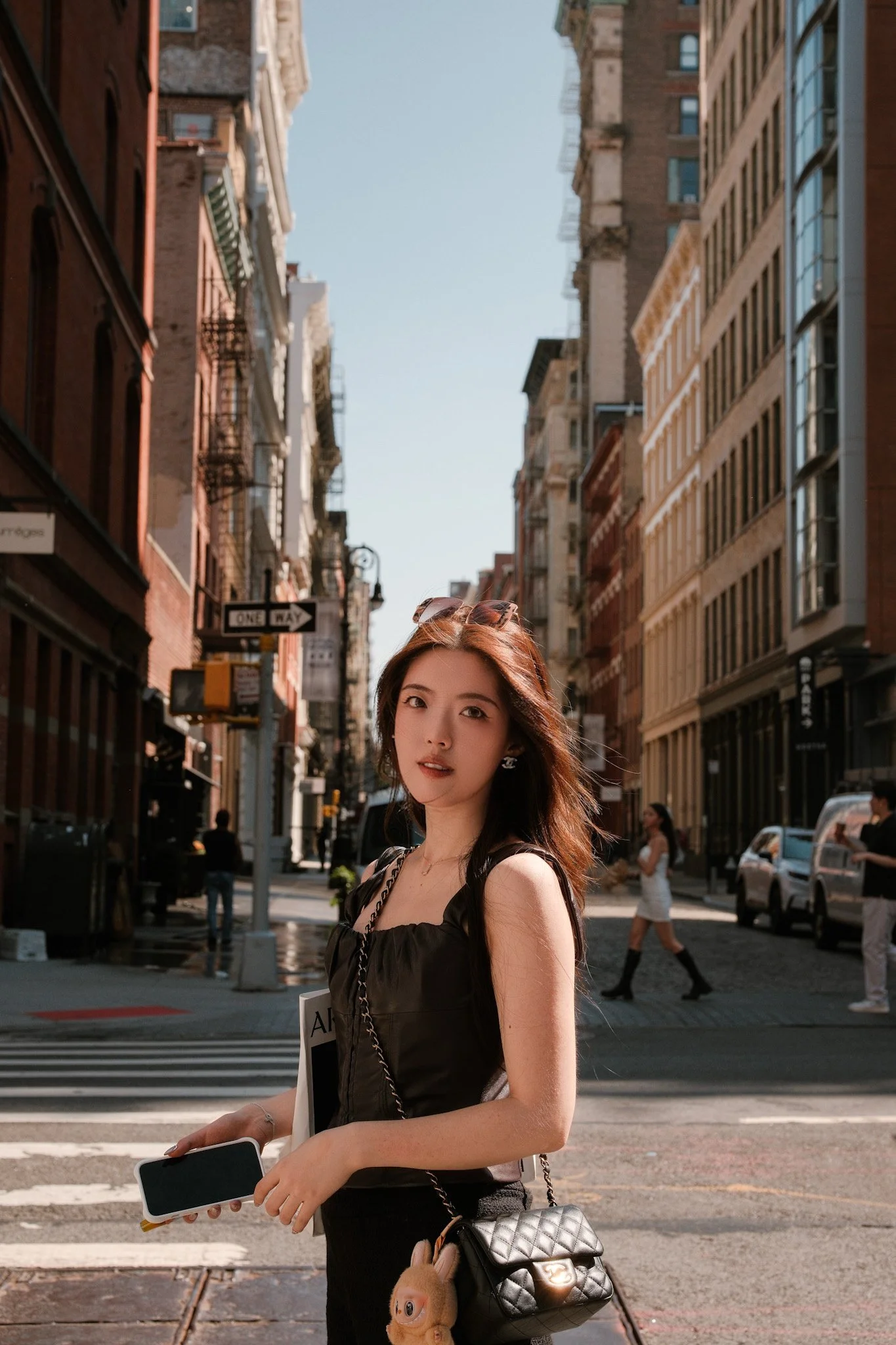 A young woman with long brown hair stands on a city street holding a smartphone and a notebook. She is dressed in a black outfit with a quilted purse hanging from her shoulder. In the background, there are tall buildings, people walking, and cars on 