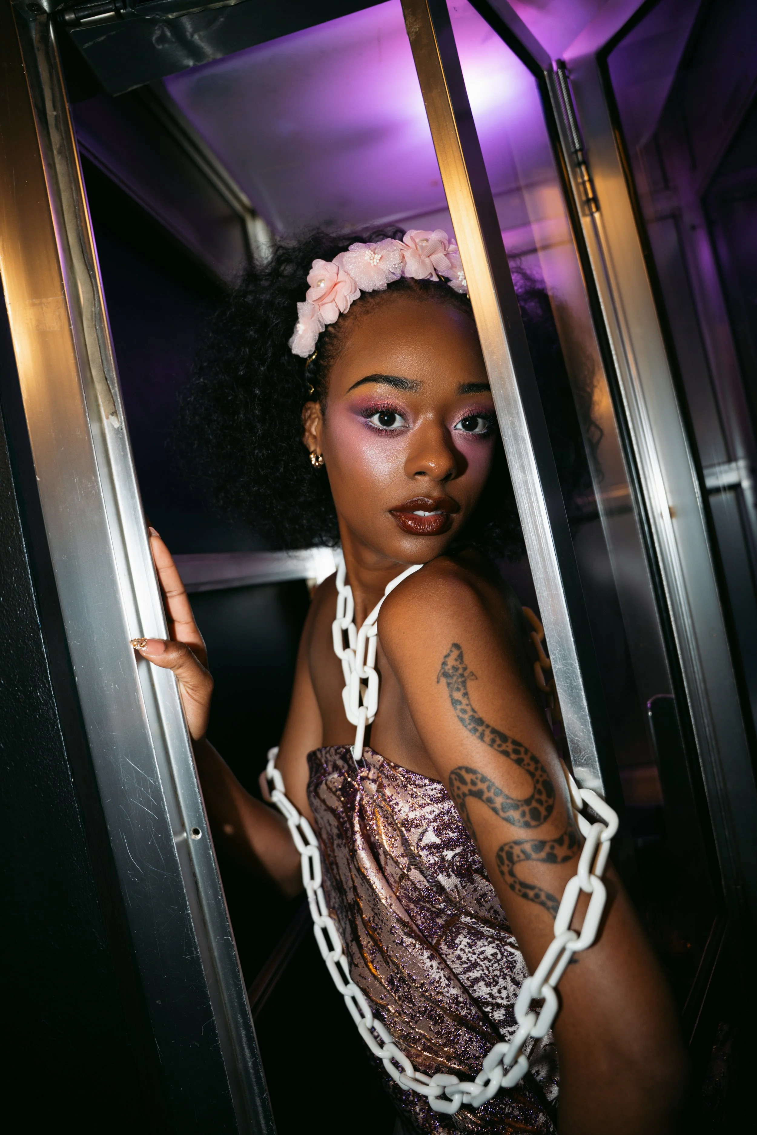 A woman with curly black hair wearing a pink floral headband and makeup, framed by metallic structures with purple lighting, holding a white chain, and displaying a leopard tattoo on her arm.