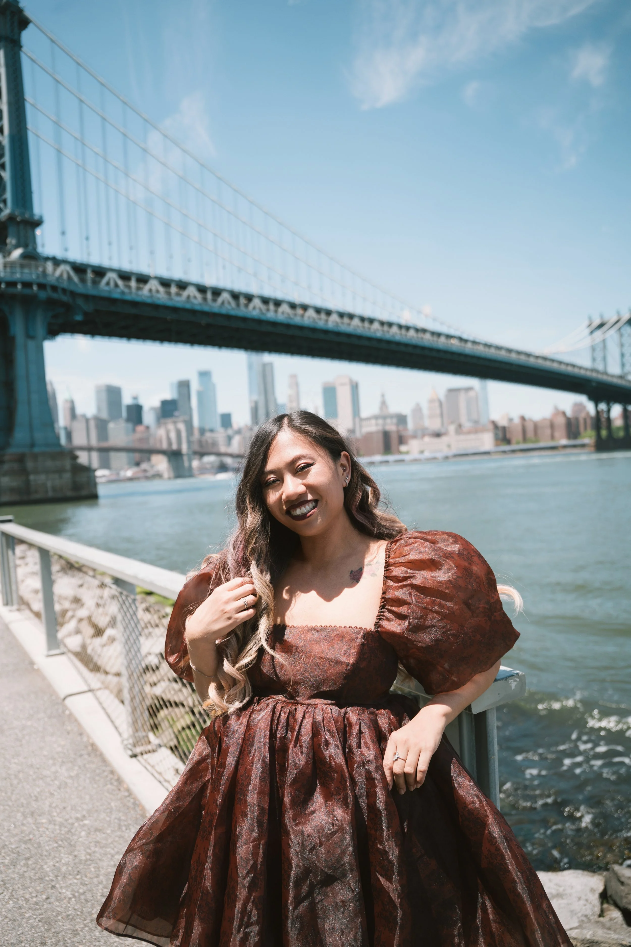 A woman with long, wavy hair and dark lipstick, dressed in a brown Gothic-style gown with puffed sleeves, standing near a waterfront with a city skyline and the Brooklyn Bridge in the background on a sunny day.