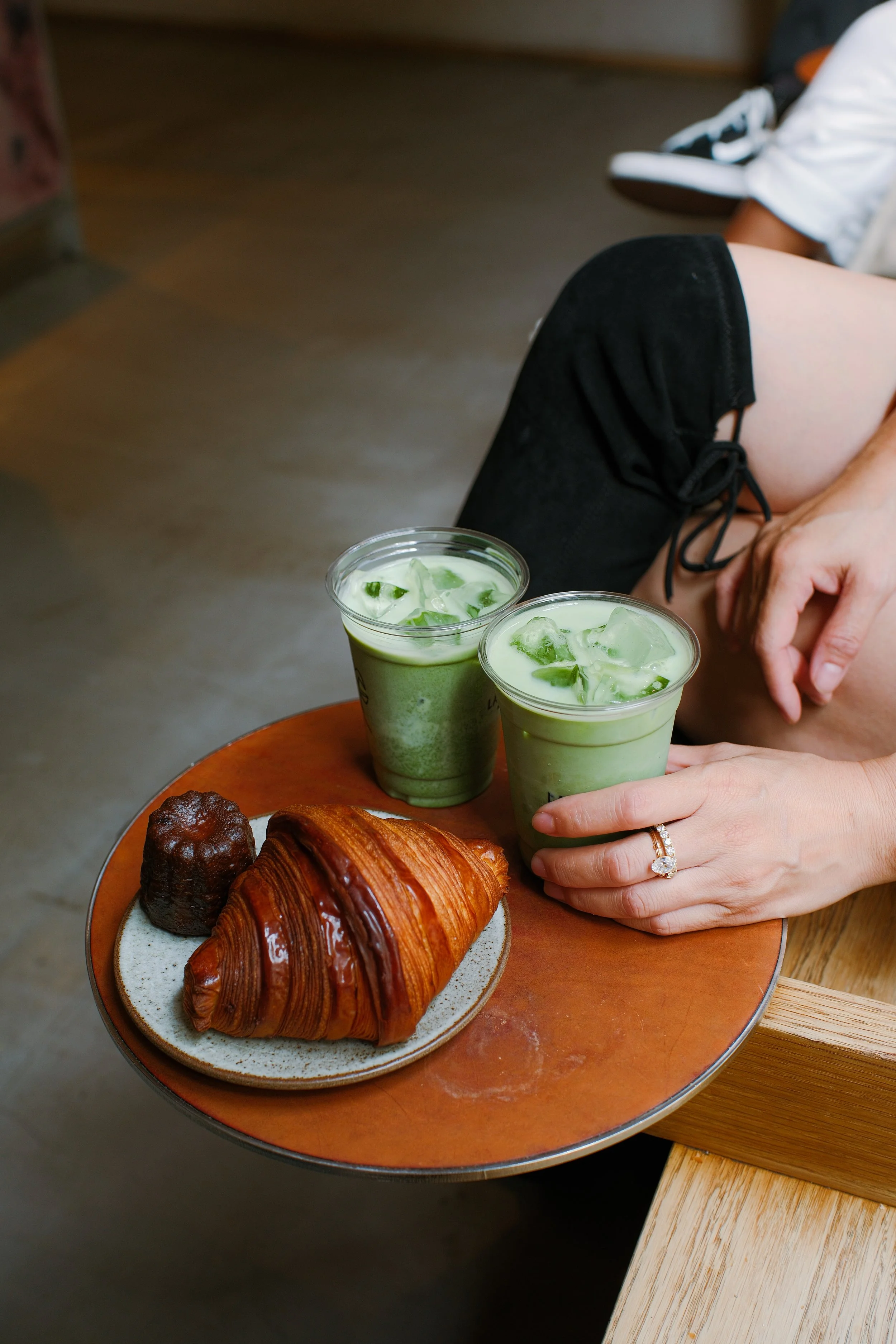 Two green drinks with ice and cucumber slices on a wooden table, along with a plate of assorted pastries including a croissant, a cube-shaped pastry, and a chocolate-covered pastry. A woman wears a ring on her finger and holds one of the drinks.