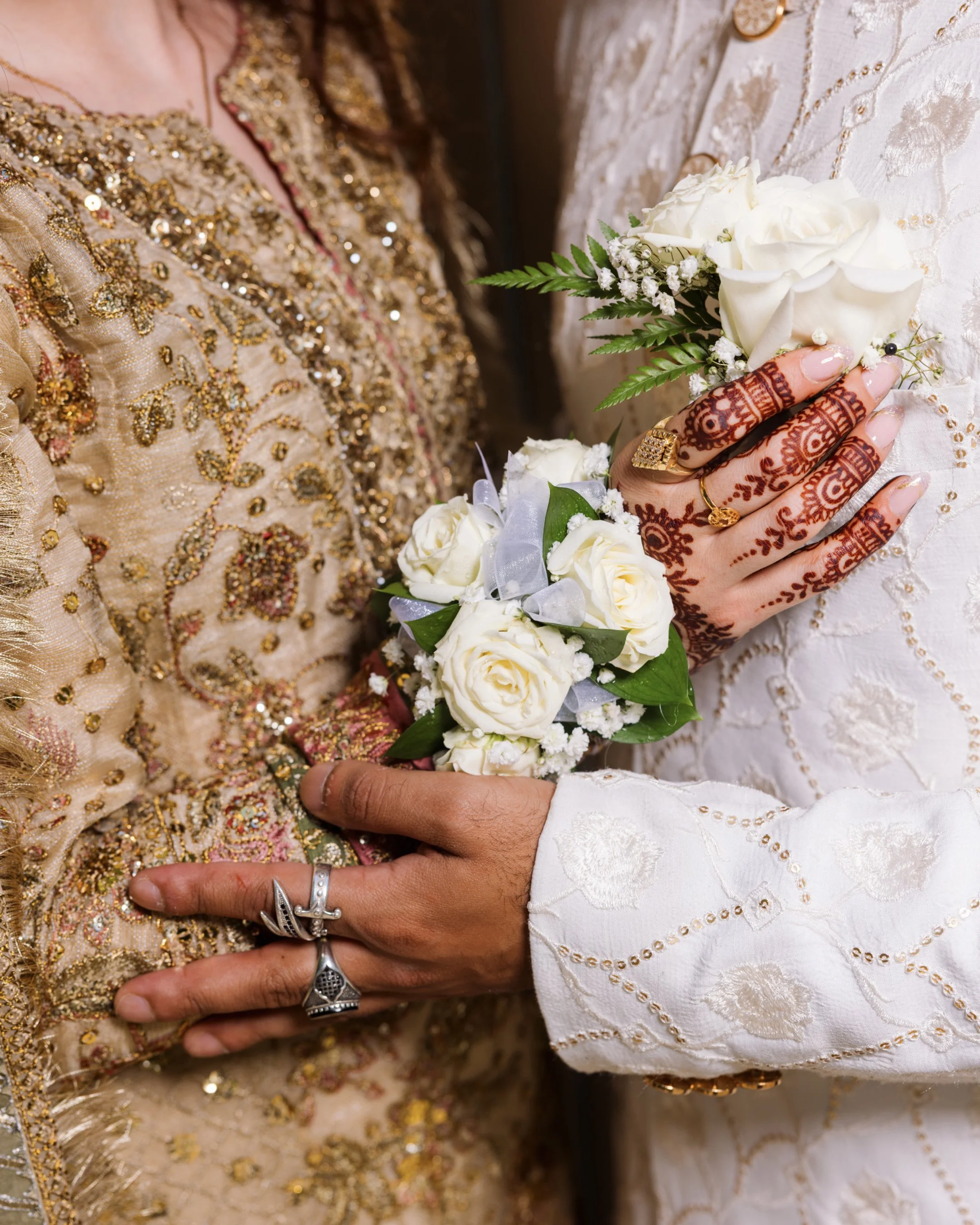 Close-up of two hands with intricate henna designs, holding a small bouquet of white roses and baby's breath, with people dressed in ornate, gold and white traditional clothing.