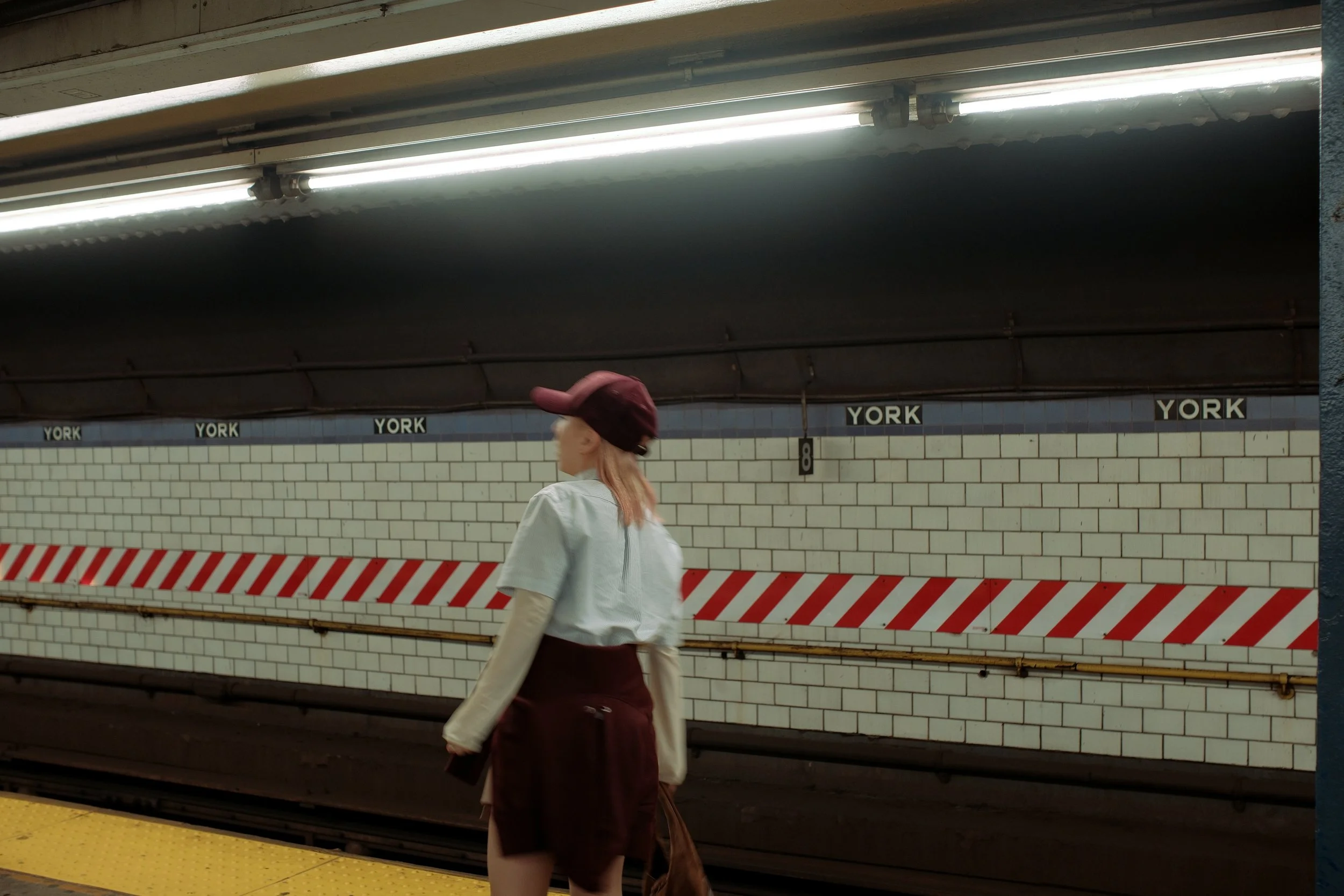 A woman with pink hair wearing a maroon beret, white top, and maroon skirt walking on the subway platform in front of a York subway station sign.