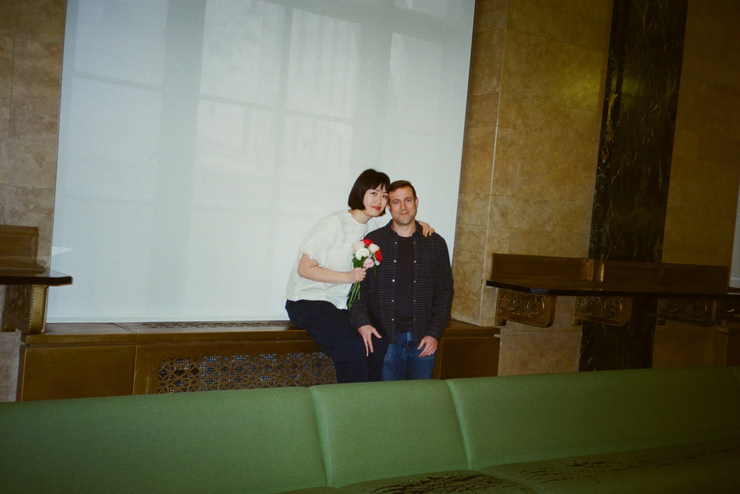 A woman holding a bouquet of flowers and a man standing beside her, both smiling, in an indoor setting with marble and wood decor.