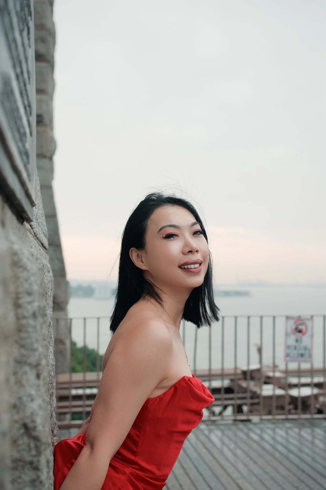 A woman with black hair wearing a red top standing outdoors on a balcony or terrace, smiling with the water and sky in the background.