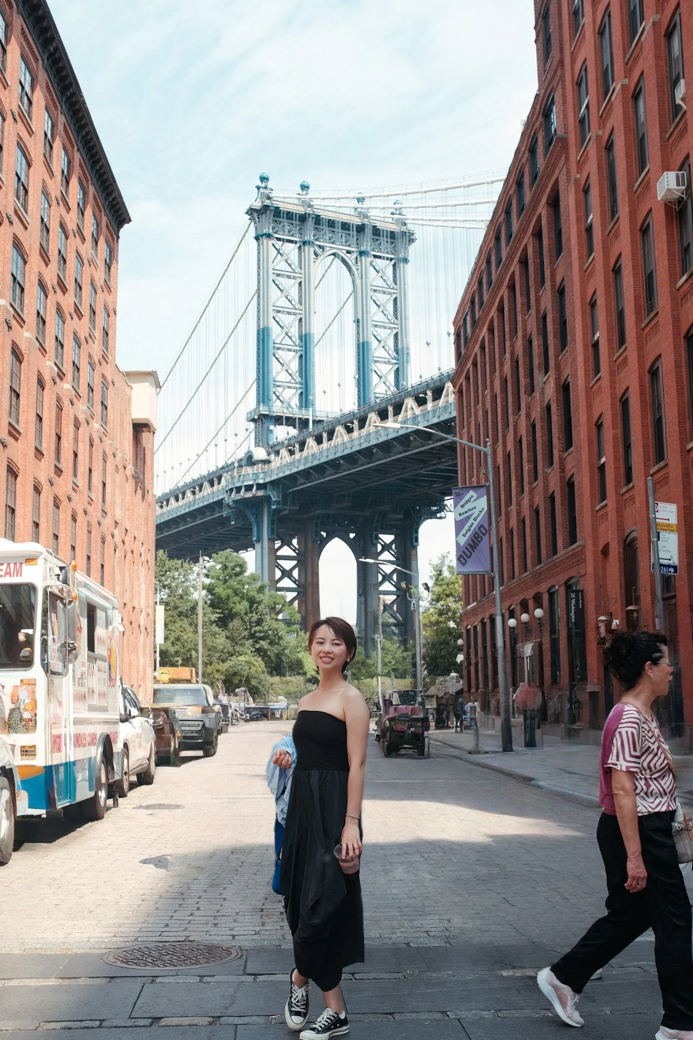 A young woman in a black dress and sneakers stands smiling in a city street with a bridge in the background. Several other people are walking by, and red brick buildings line the street.