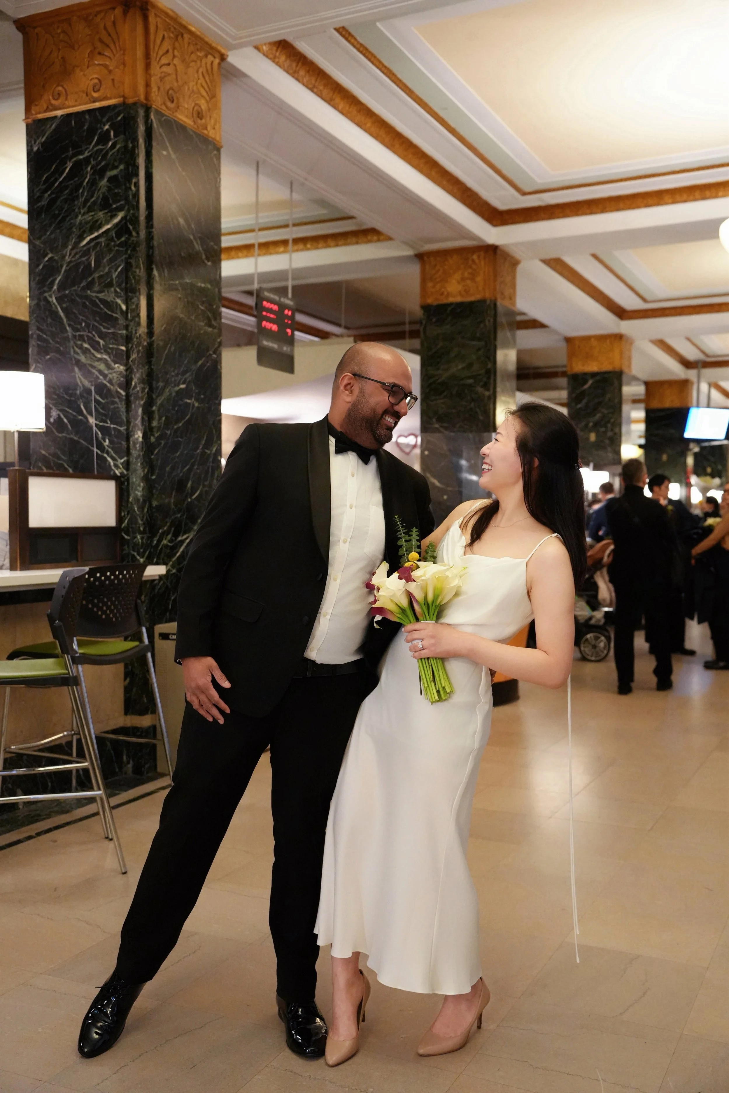 A man and woman dressed in formal attire, smiling and looking at each other in a wedding or celebration setting. The man wears a black tuxedo with a bow tie, and the woman wears a white gown, holding a bouquet of flowers. They are inside a decorated 