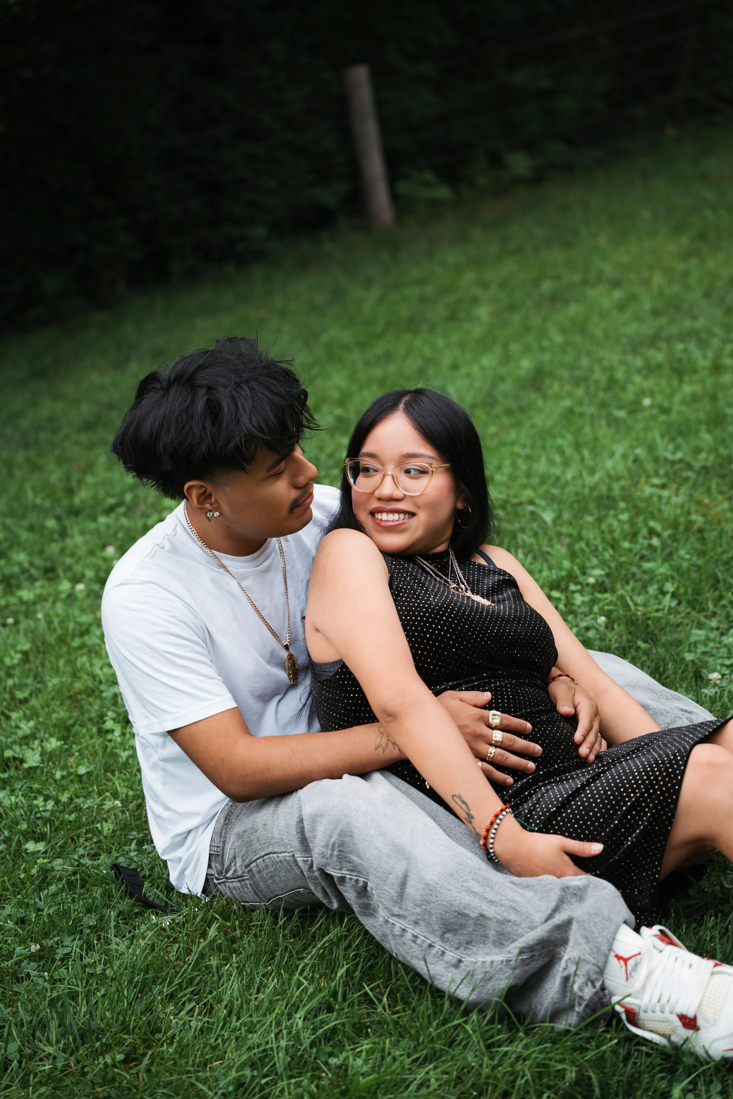 A young man and woman sitting on a grassy field, embracing each other and smiling. The woman wears glasses and a black dress with gold dots, while the man wears a white t-shirt and gray jeans.