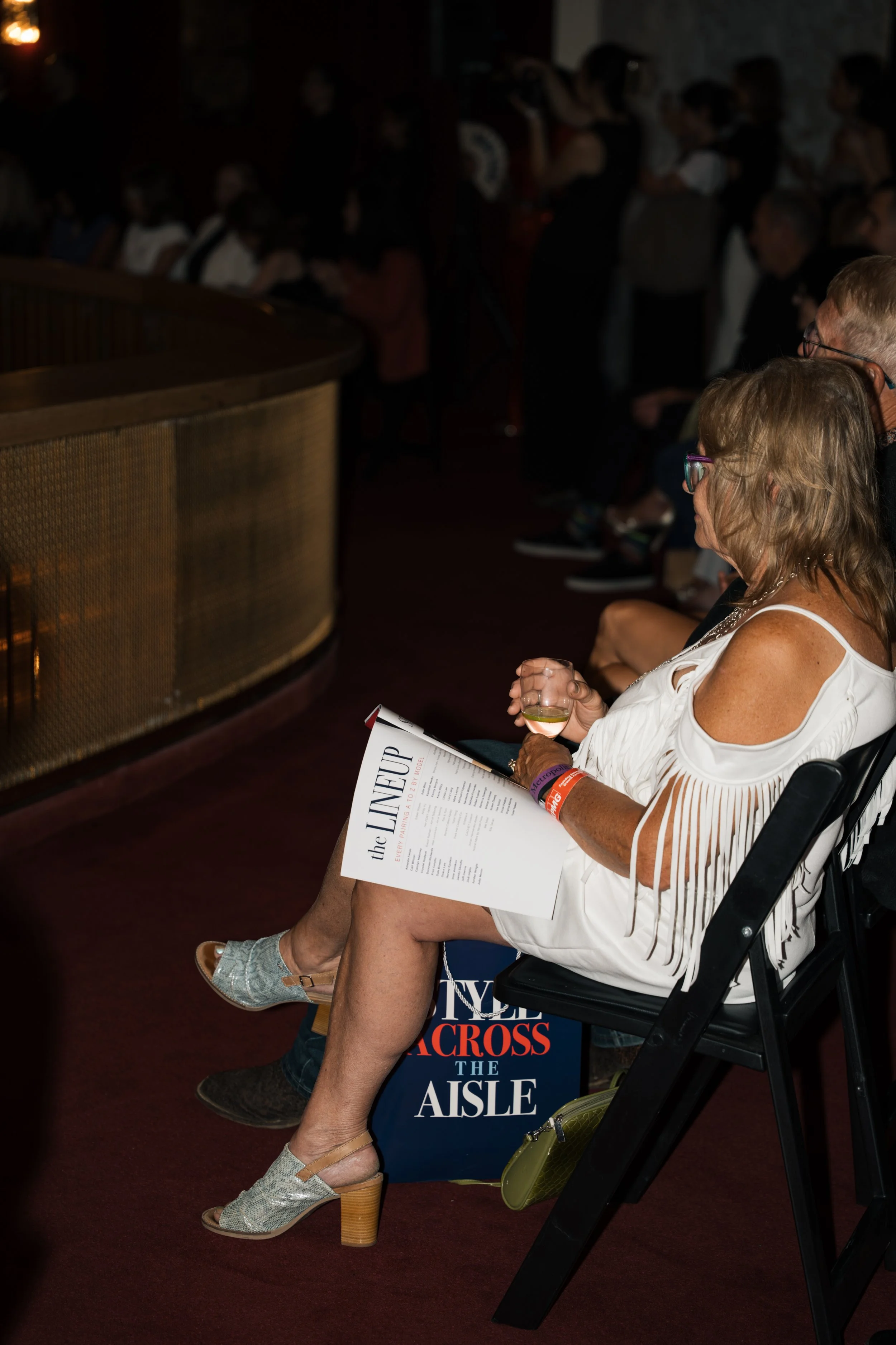 A woman in a white dress sitting in a black folding chair, holding a glass of white wine, reading a magazine, with a protest sign that says "the LINKSUP the CROSS the AISLE" in front of her at an indoor event.