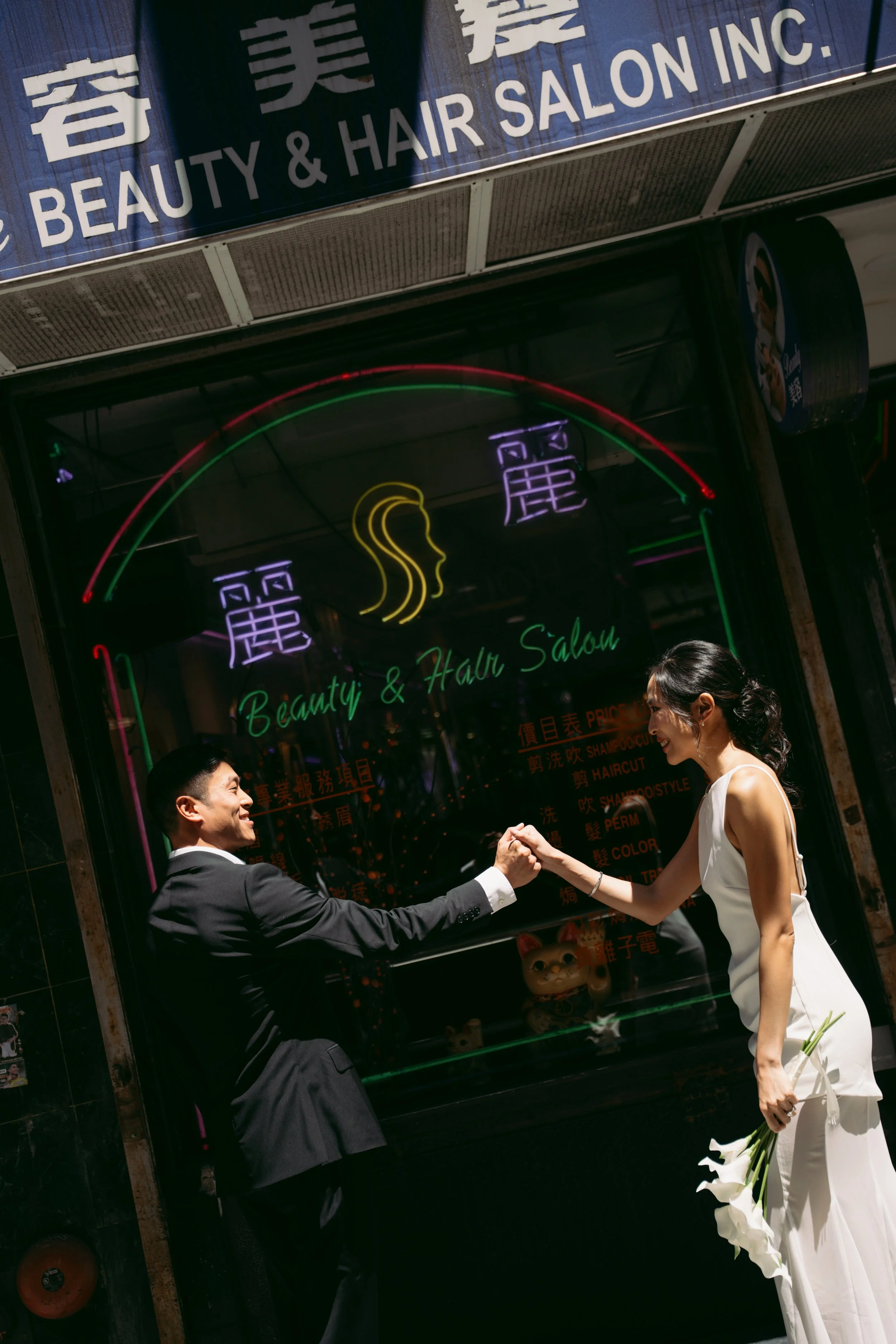 A man in a suit and a woman in a white dress shake hands outside a beauty and hair salon, both smiling, with the woman holding a bouquet of white calla lilies.