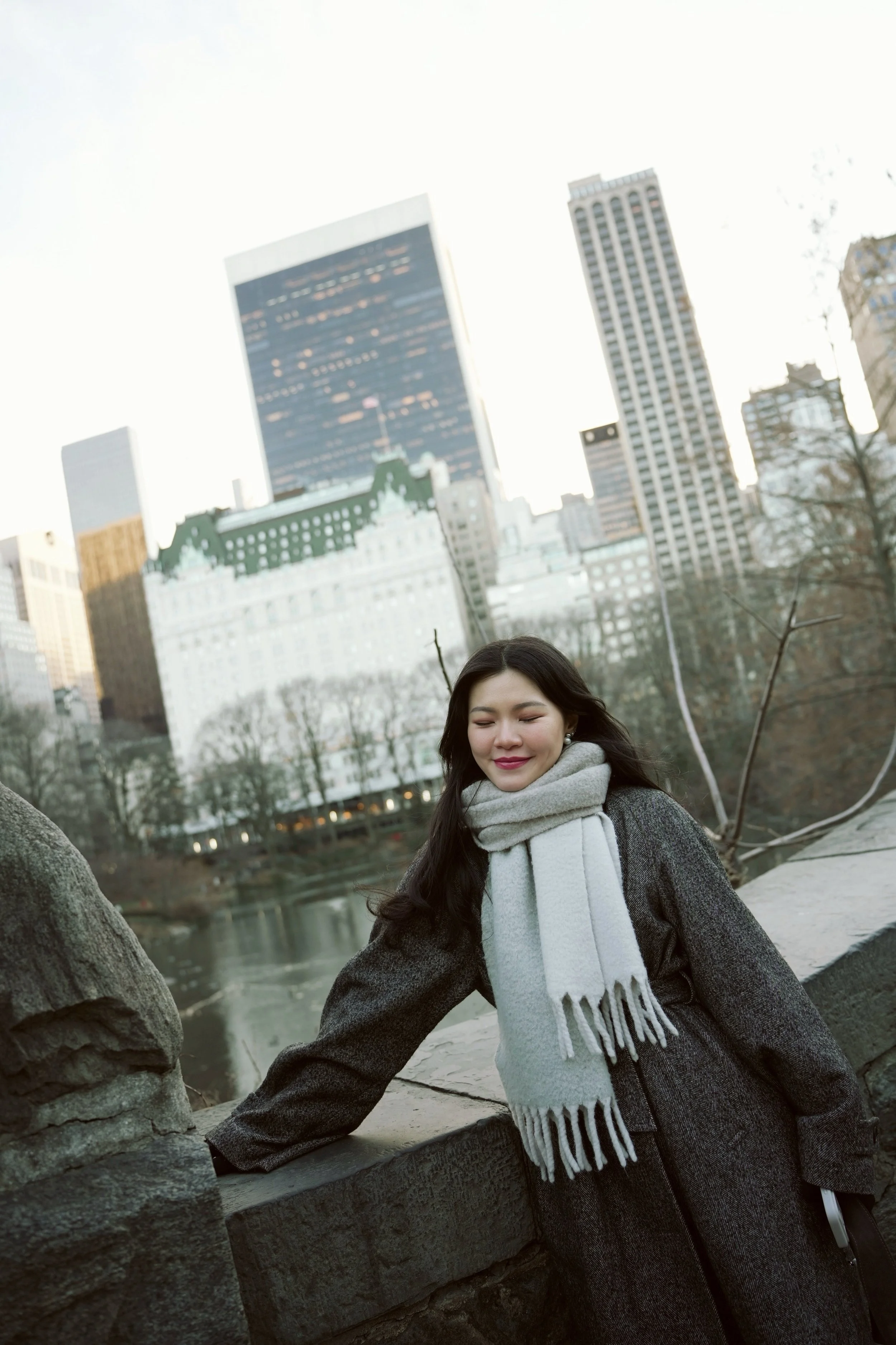 A woman with long dark hair, wearing a grey coat and a white scarf, standing outdoors near a stone railing with a city skyline of tall buildings and a river in the background on a clear day.