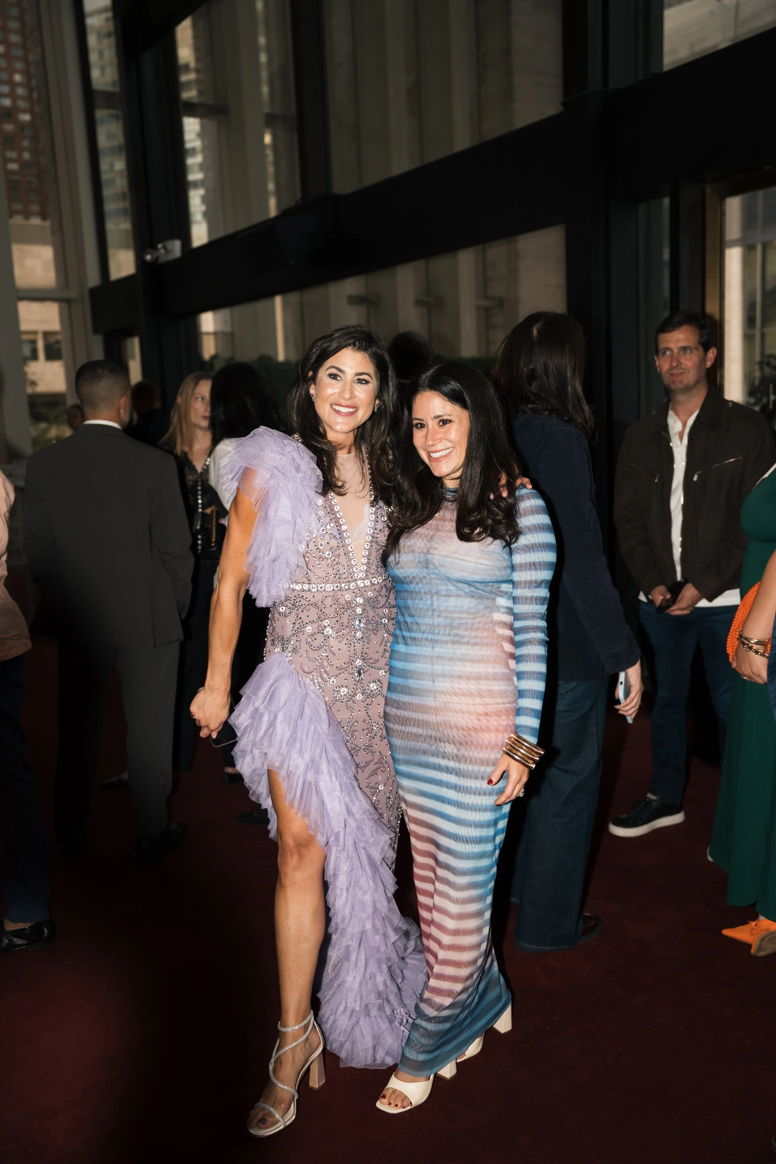 Two women are smiling and posing for a photo at an indoor event, with one in a purple ruffled dress and the other in a multi-colored striped dress.