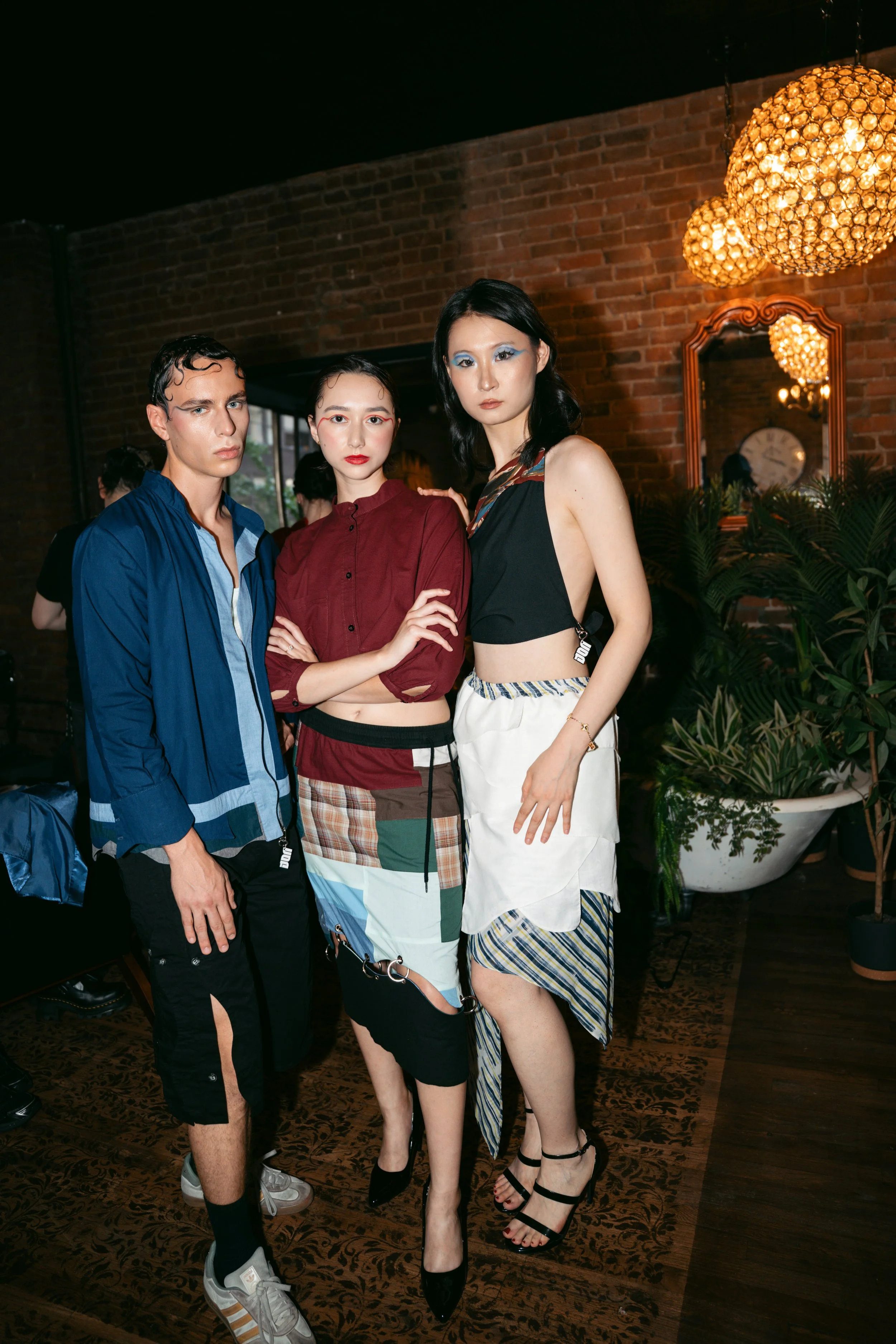 Three models with bold makeup and fashion-forward outfits posing indoors with a brick wall, plants, and hanging lights in the background.