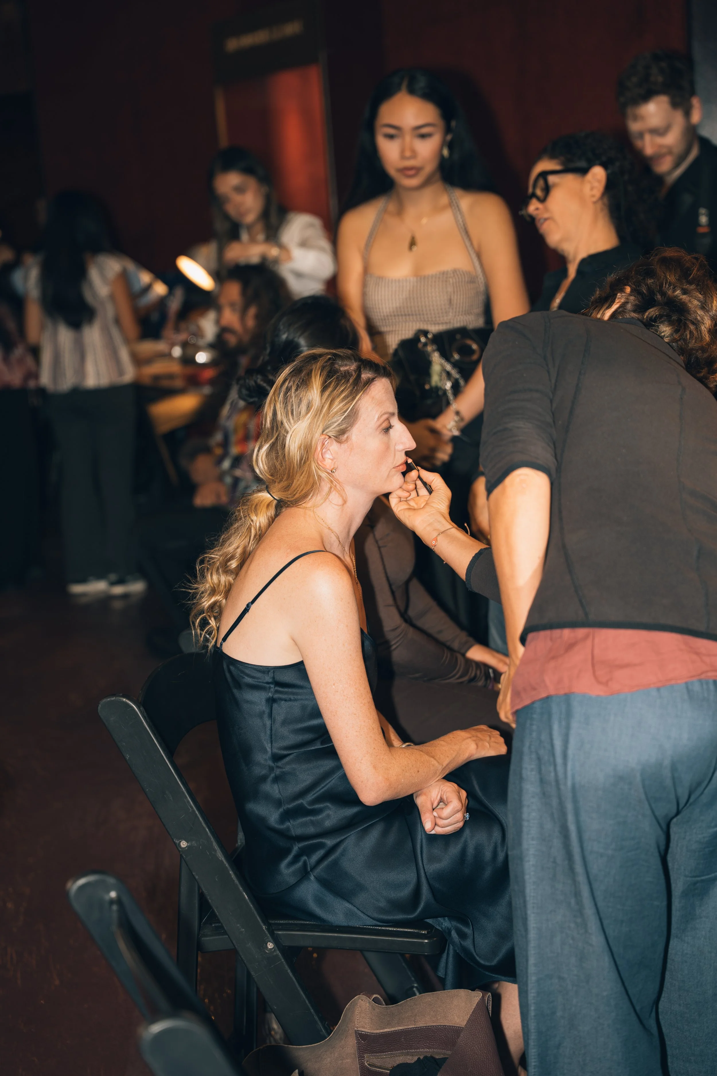 A woman with long blonde hair sitting in a chair, having her makeup done by a makeup artist. The woman is wearing a black satin dress and the makeup artist is standing and working on her lips. People are in the background, some talking and others wor