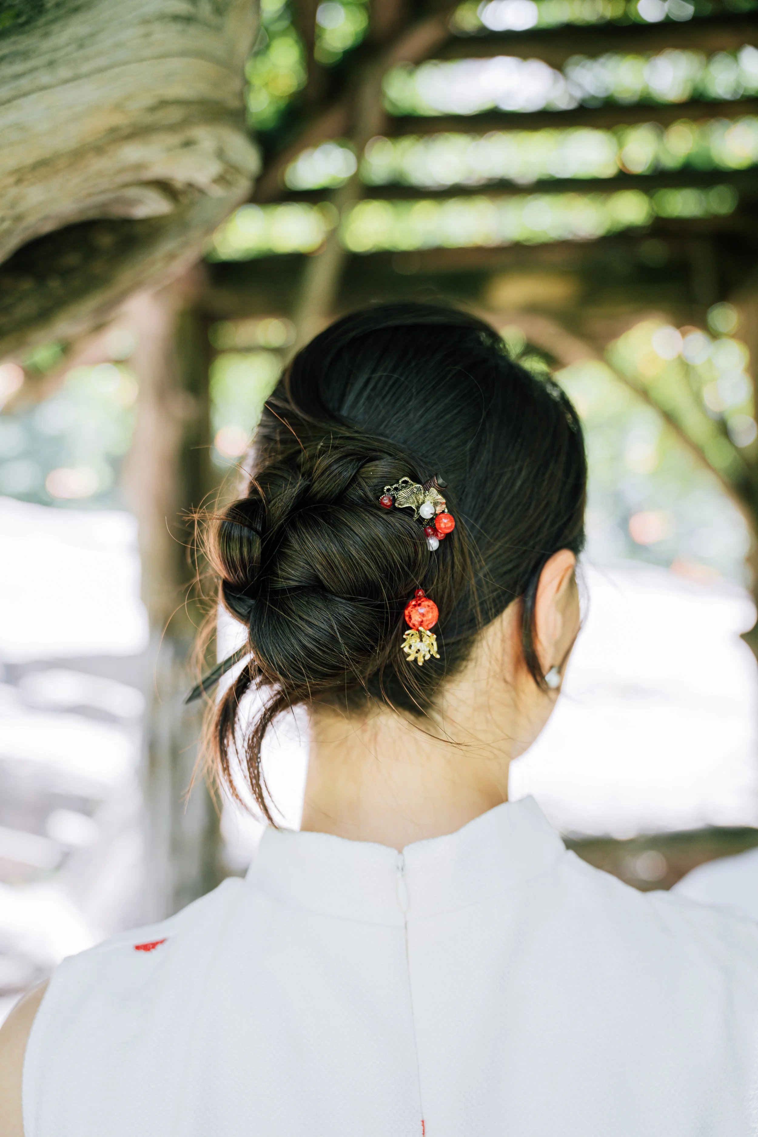Back view of a woman with dark hair styled in an elegant updo, decorated with red and white floral hair accessories, wearing a white garment, outdoors with blurred greenery and wooden structures in the background.