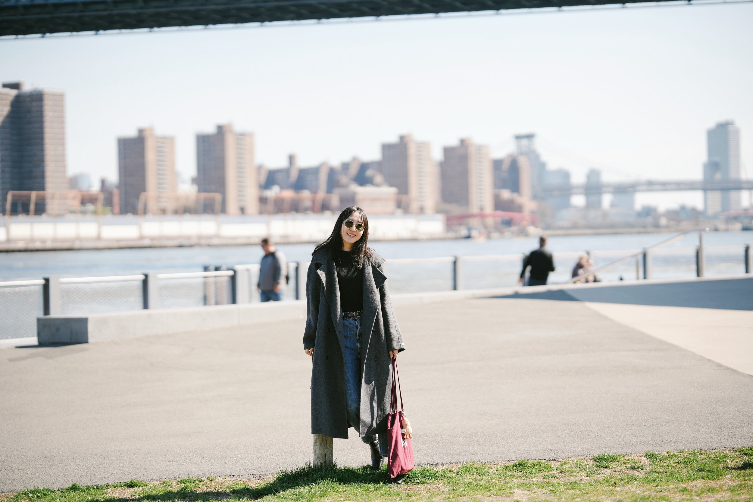 A woman with long dark hair and sunglasses, wearing a long dark coat and blue jeans, standing outdoors next to a wooden post by the waterfront. She is smiling and carries a pink bag. In the background, there are blurred city buildings and a bridge ac