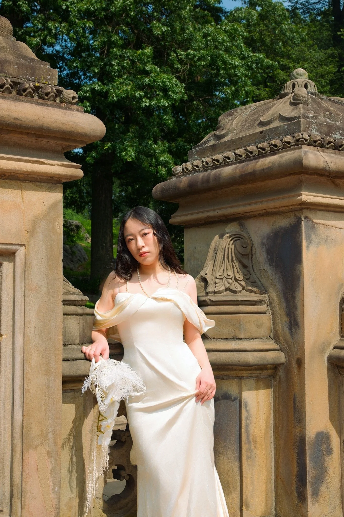 A woman in a white dress holding a lace parasol standing between two stone columns with ornate carvings, with green trees and blue sky in the background.