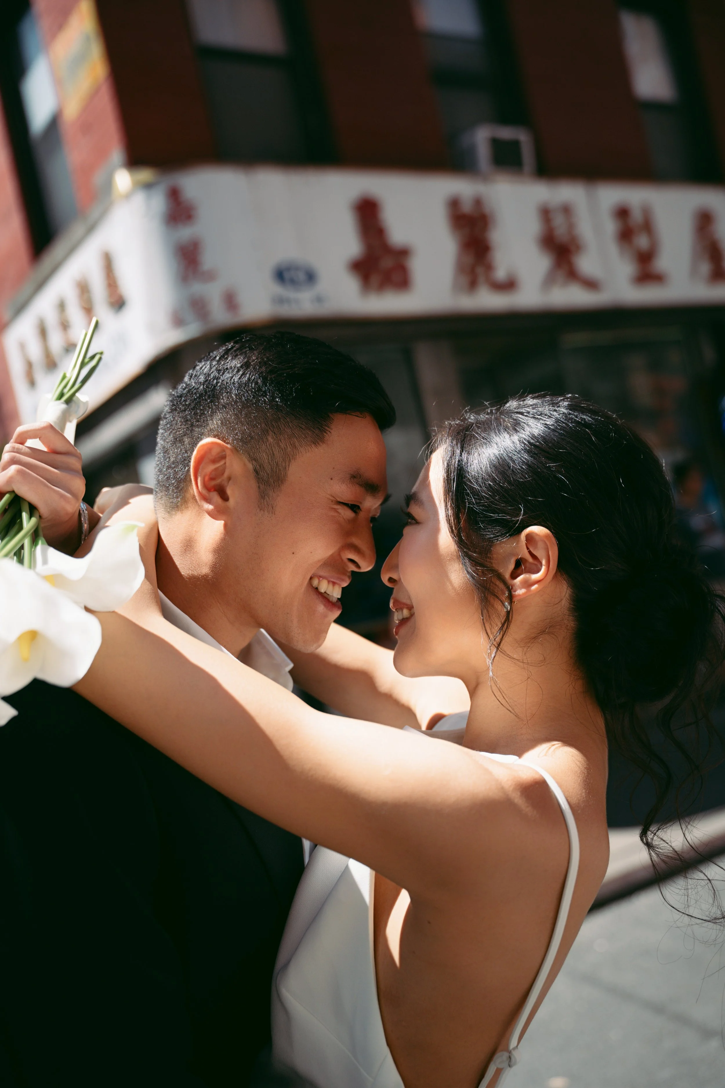 A happy couple, man and woman, embracing and touching foreheads, smiling at each other outdoors, with a building and signs in the background.