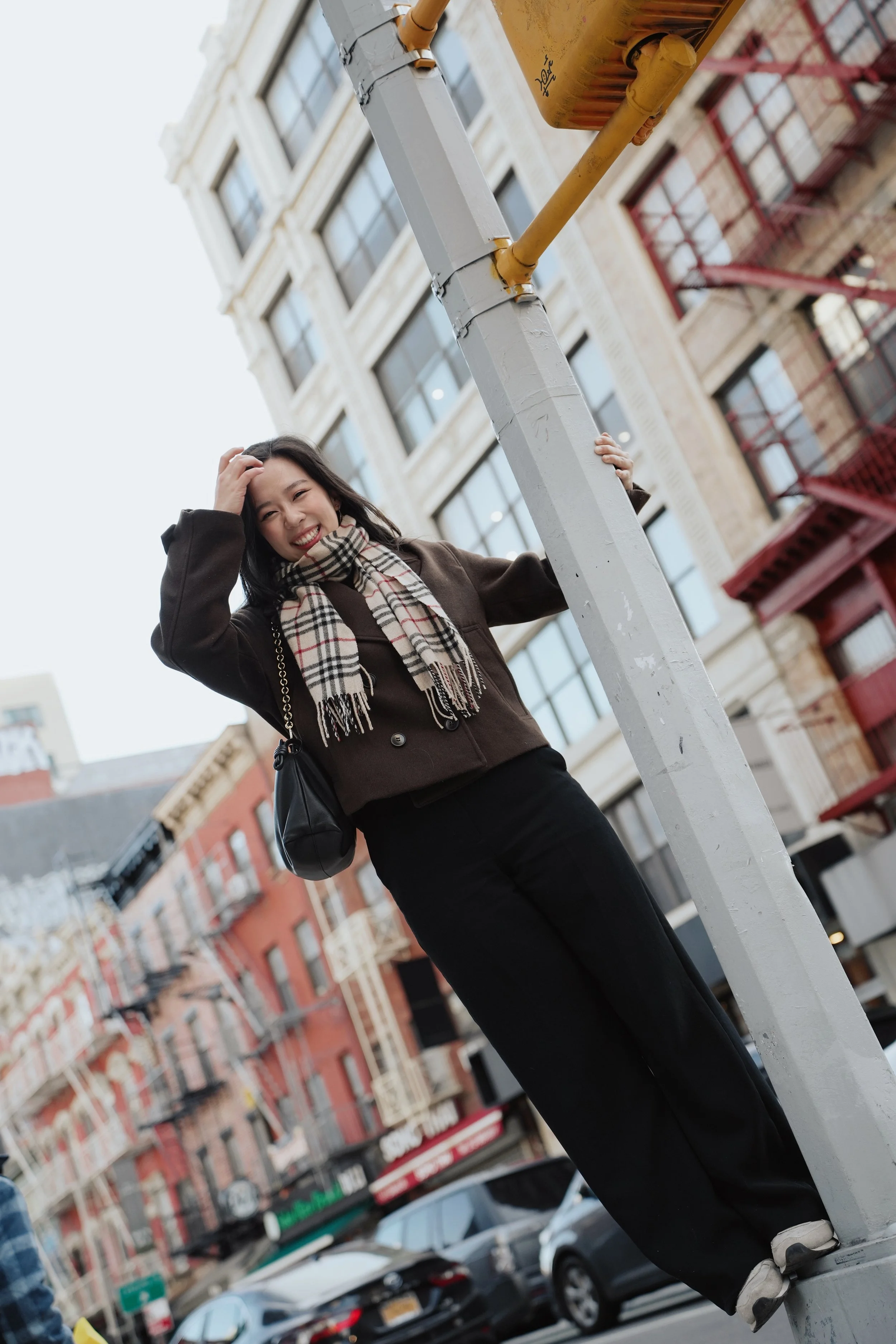 Young woman smiling, standing outdoors in front of a city street with parked cars and multi-story brick buildings, holding onto a streetlight pole.